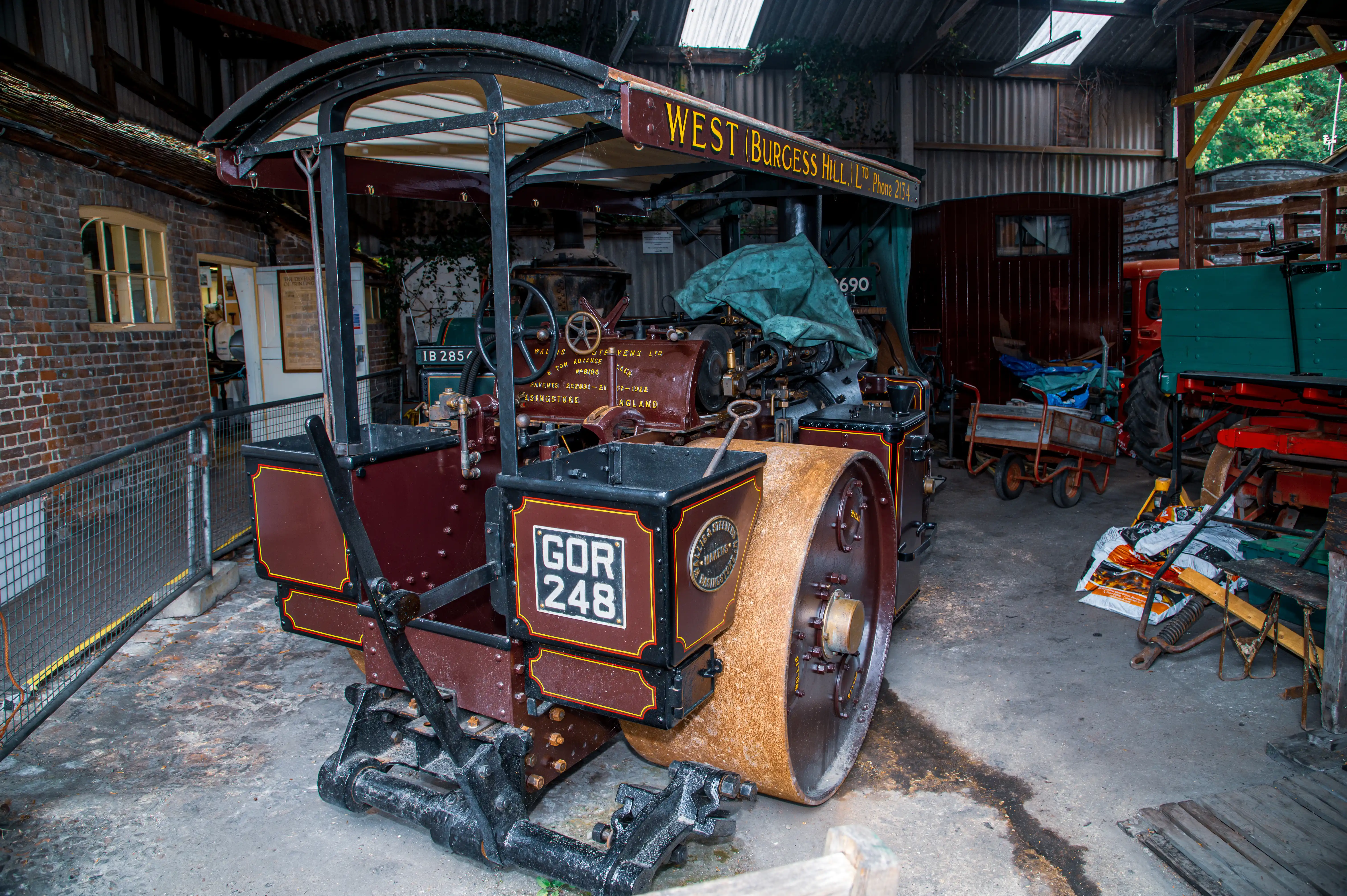 Maroon and black steam road roller with gold trim and registration GOR 248 inside a corrugated shed with other vintage machinery.