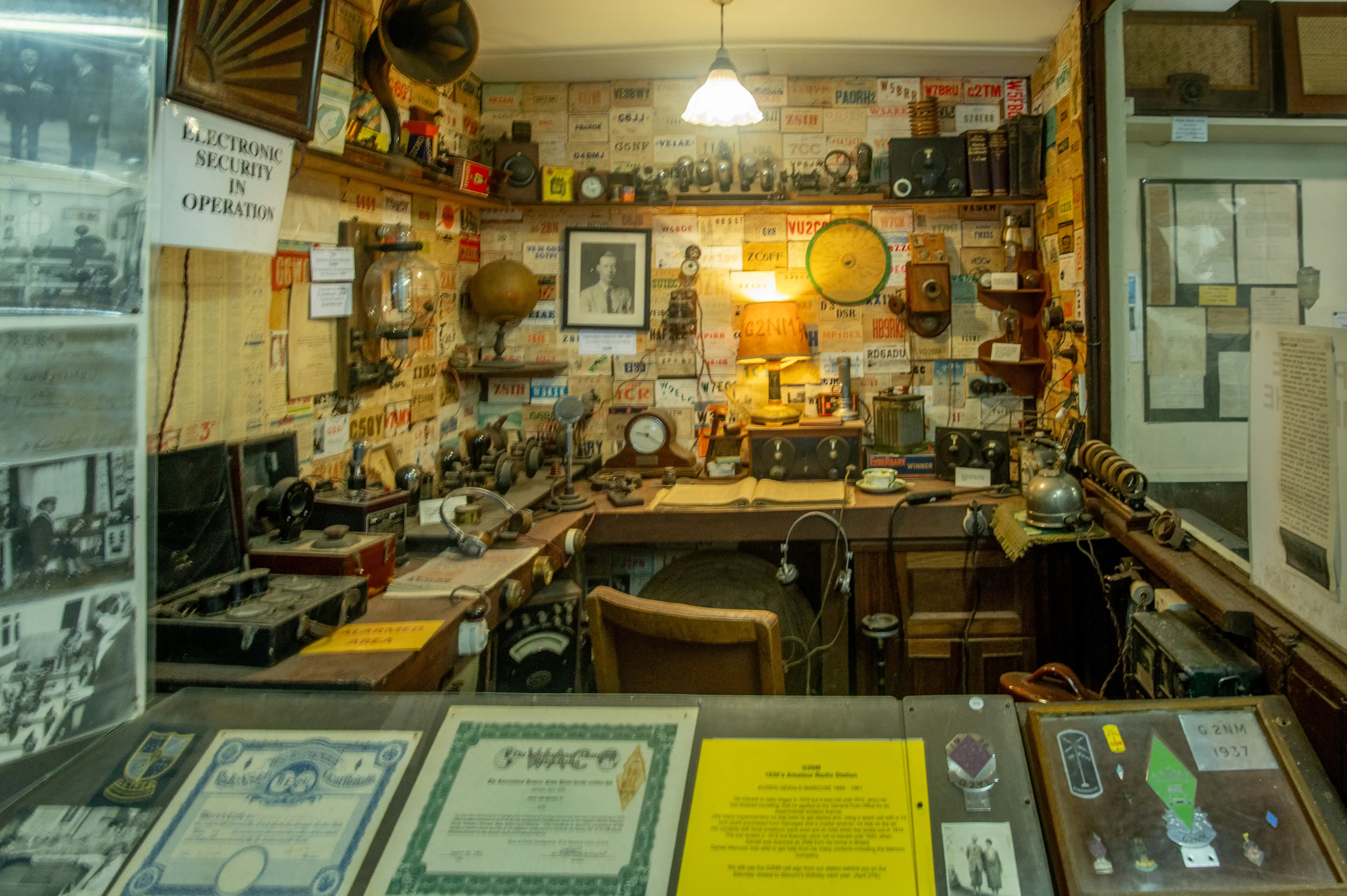 Room filled with vintage radios, transmitters, receivers, headphones, microphones, gauges, and walls covered in amateur radio call signs and QSL cards.