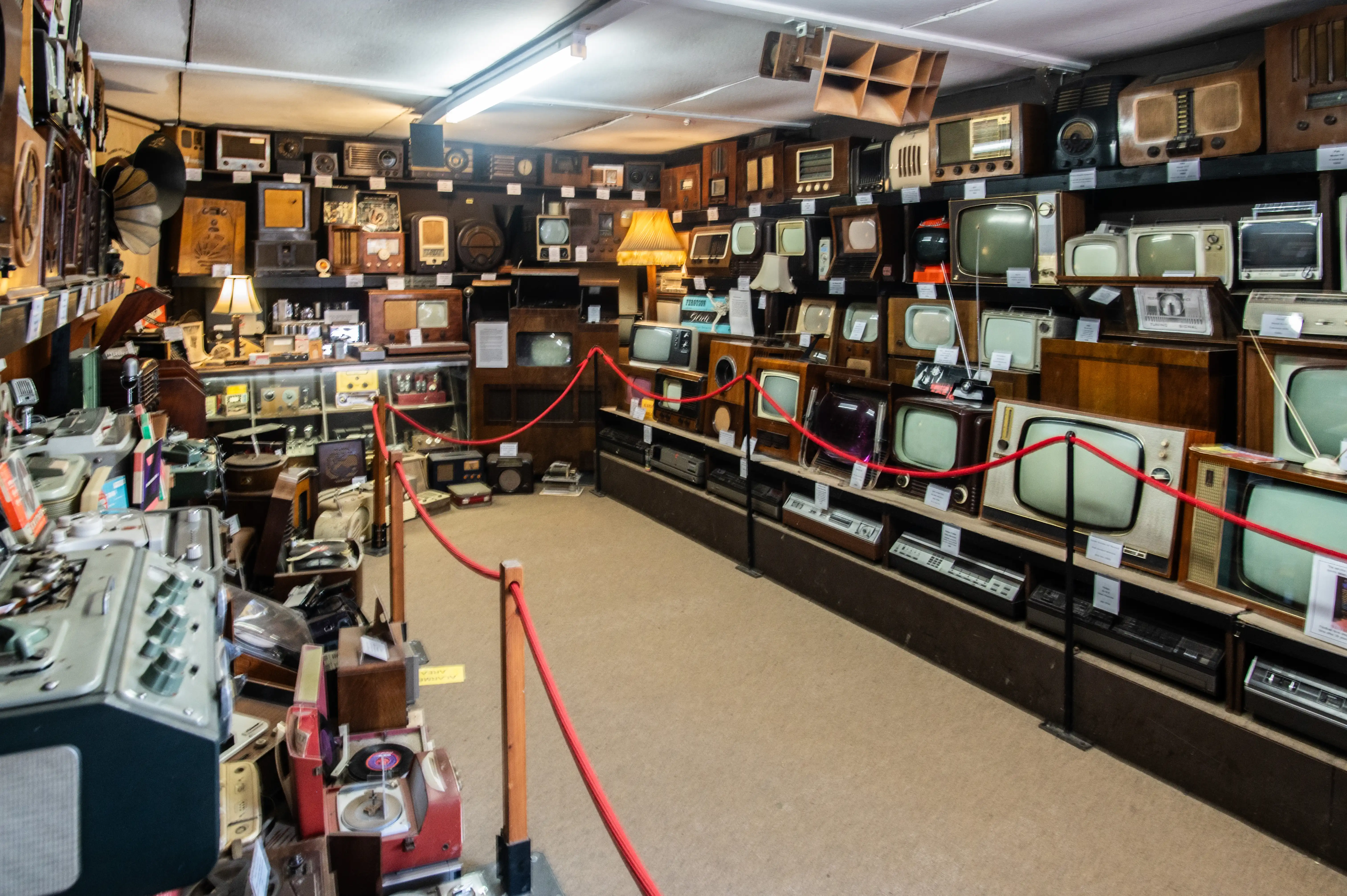 Room filled with vintage televisions, radios, amplifiers, and record players arranged on shelves with a red rope barrier and warm lighting.