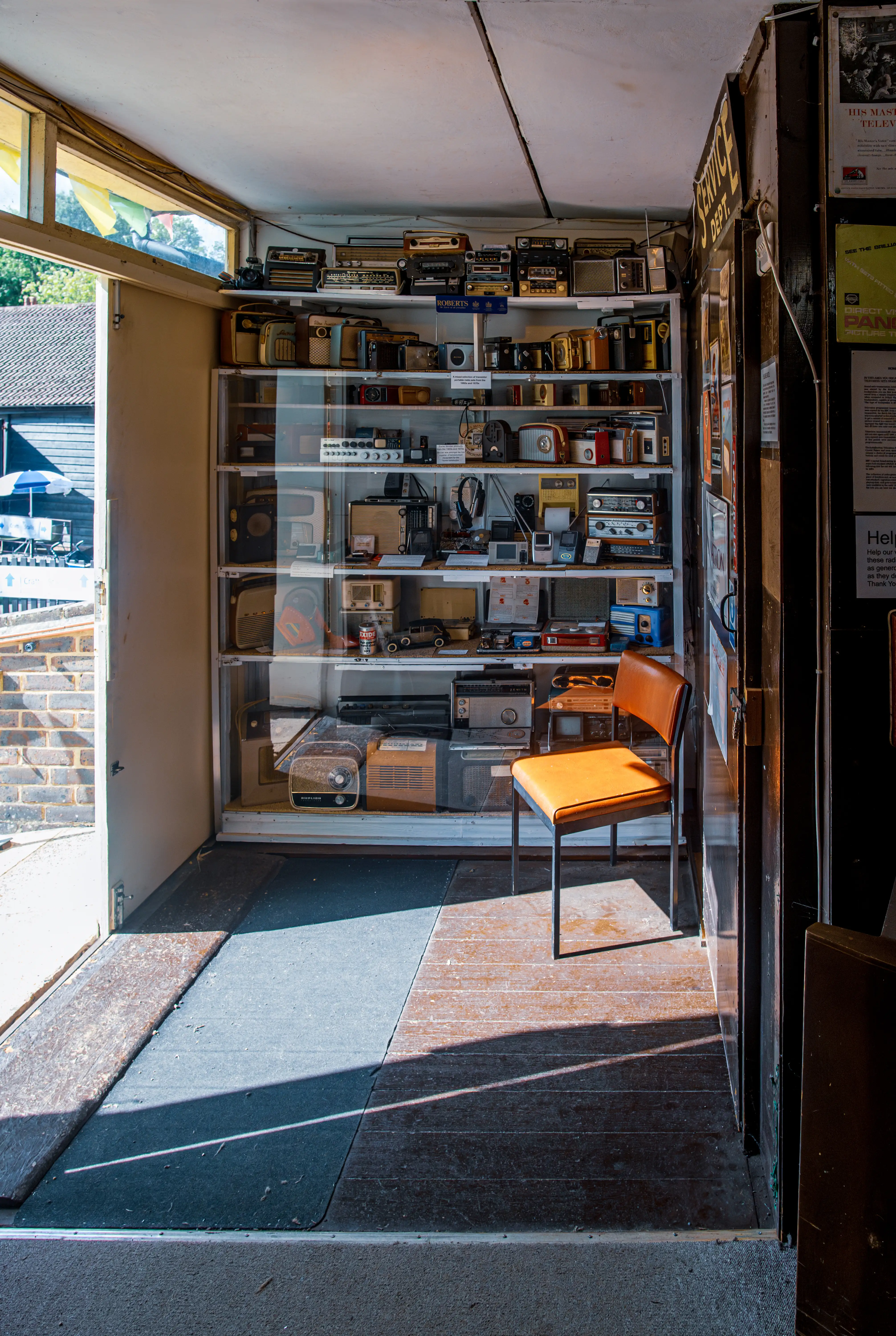 Shelves of vintage radios and cassette players with an orange chair in a small room lit by natural light from an open doorway.