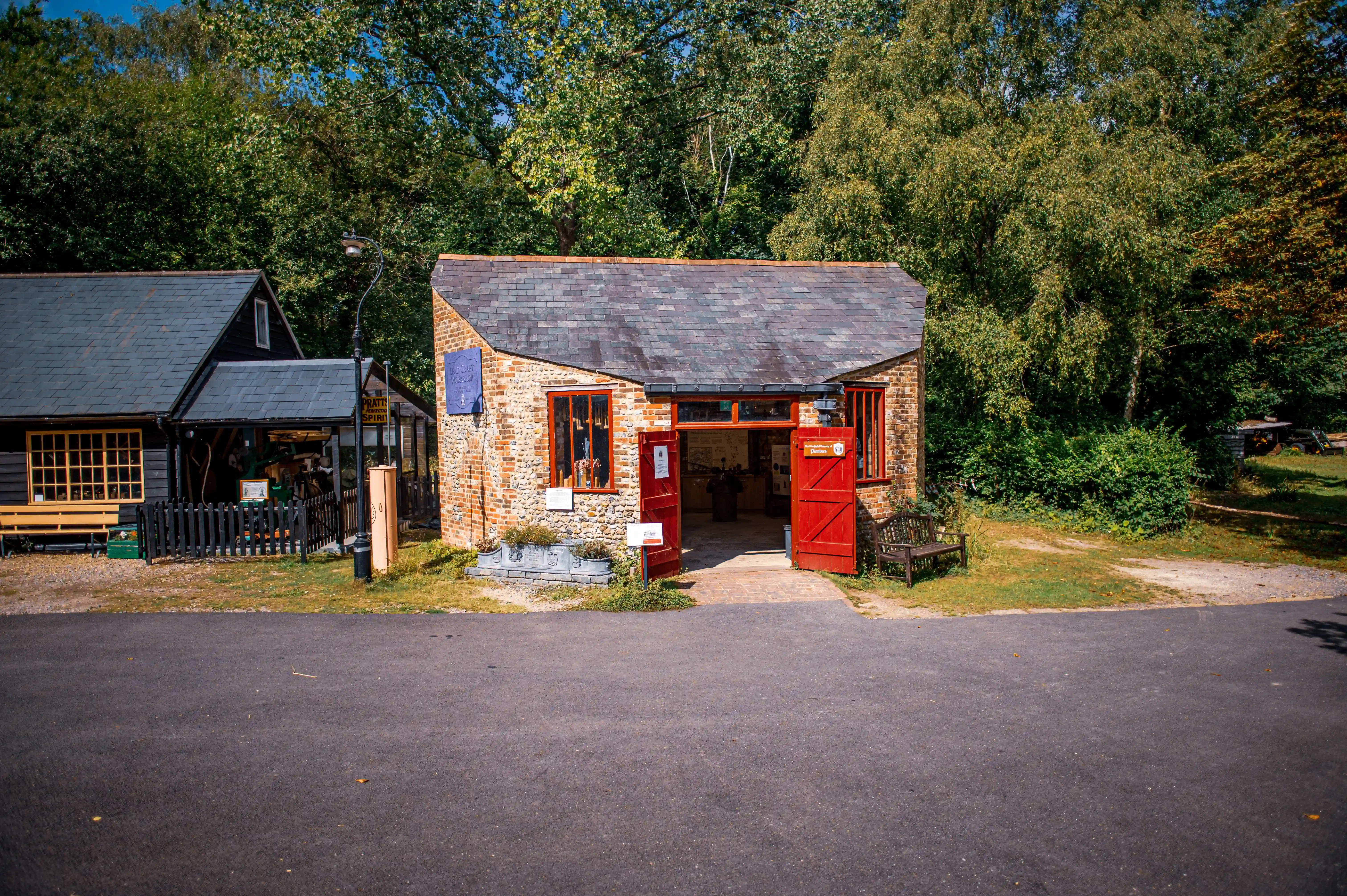 An exterior view of a brick blacksmith's forge with red double doors open, situated in a woodland setting. To the left, a dark timber building with a sloped roof is visible.