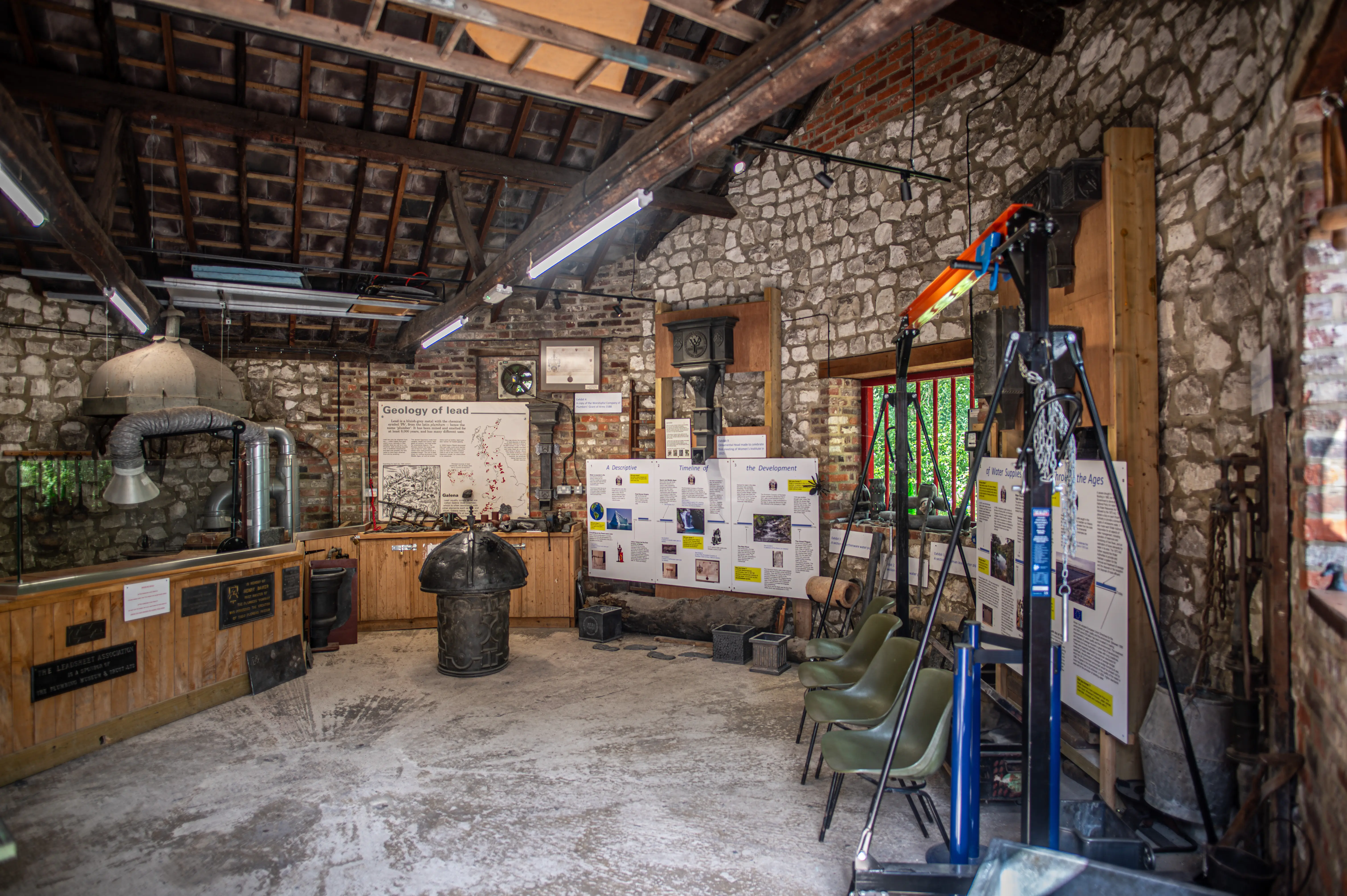 A rustic museum exhibit in a stone-walled room with wooden beams and a thatched roof. The exhibit includes informational posters, historical artifacts, mining equipment, a large metal cauldron, and a tripod setup with tools. Modern lighting illuminates the space