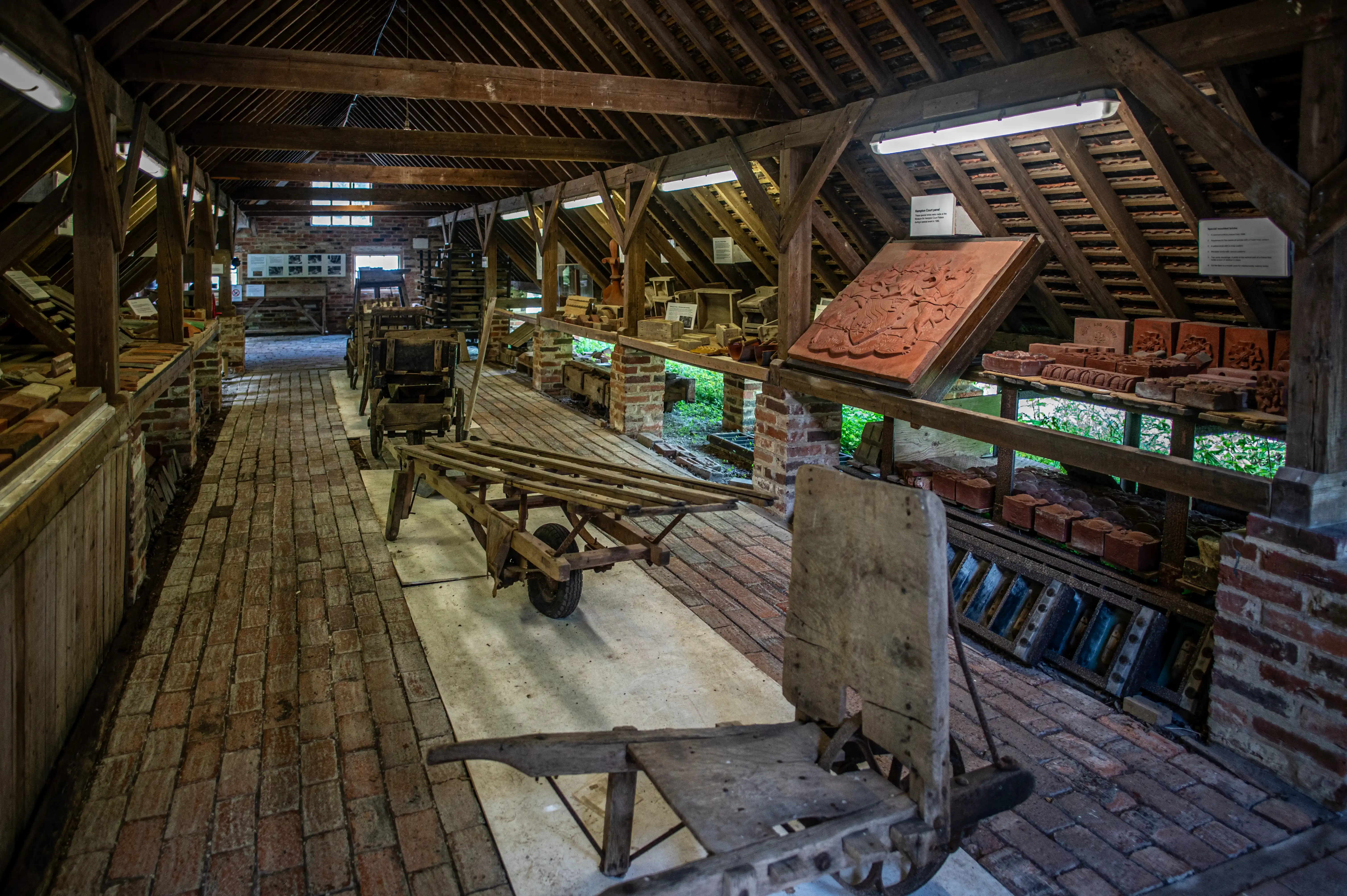Interior of a rustic brickworks museum, showing brick floors, wooden beams, shelves displaying various handmade bricks, antique trolleys, and informative signs.