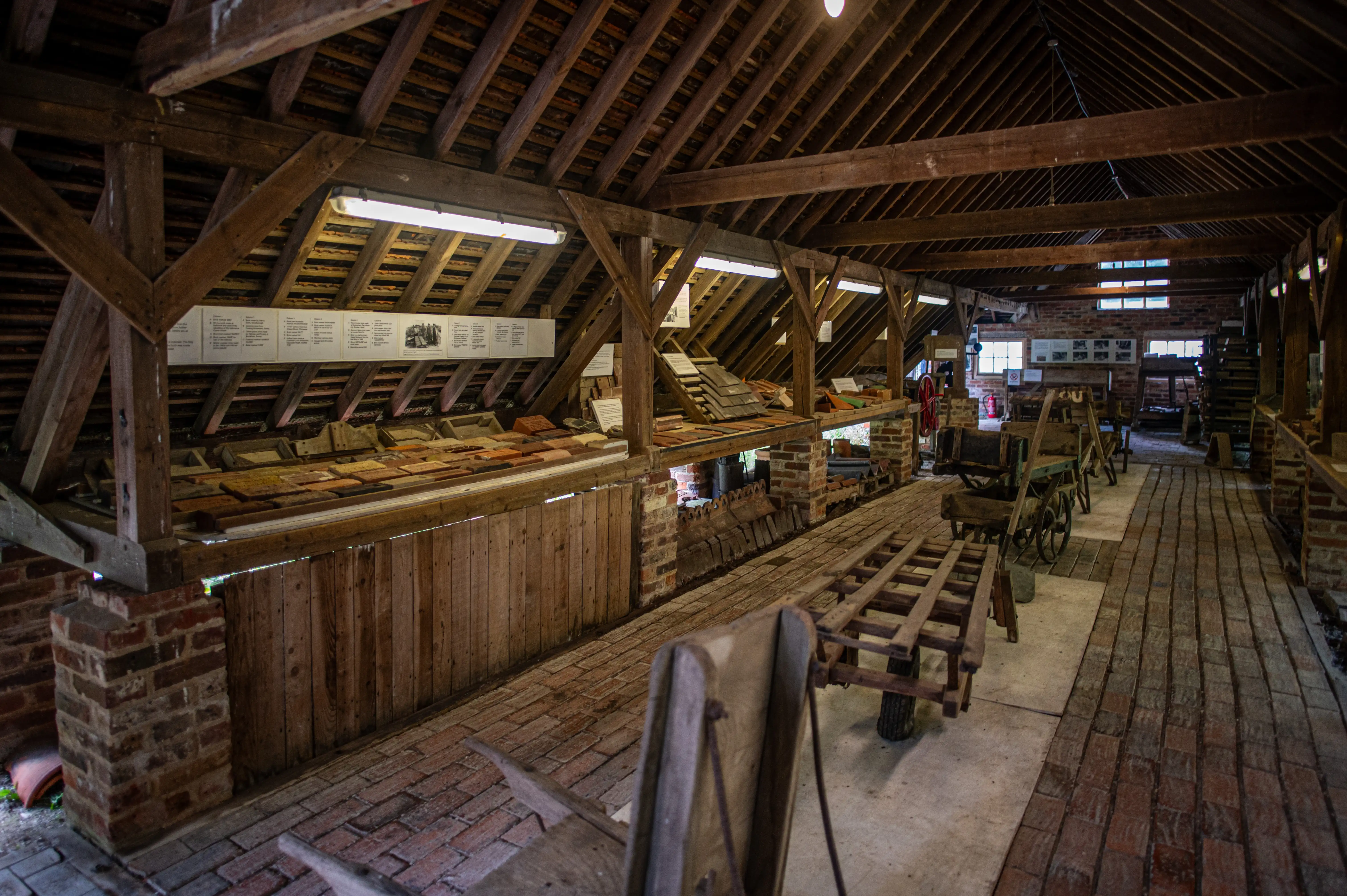 Inside a rustic museum building with a wooden beamed ceiling, display shelves filled with bricks and tiles, and old wooden trolleys on a brick floor.