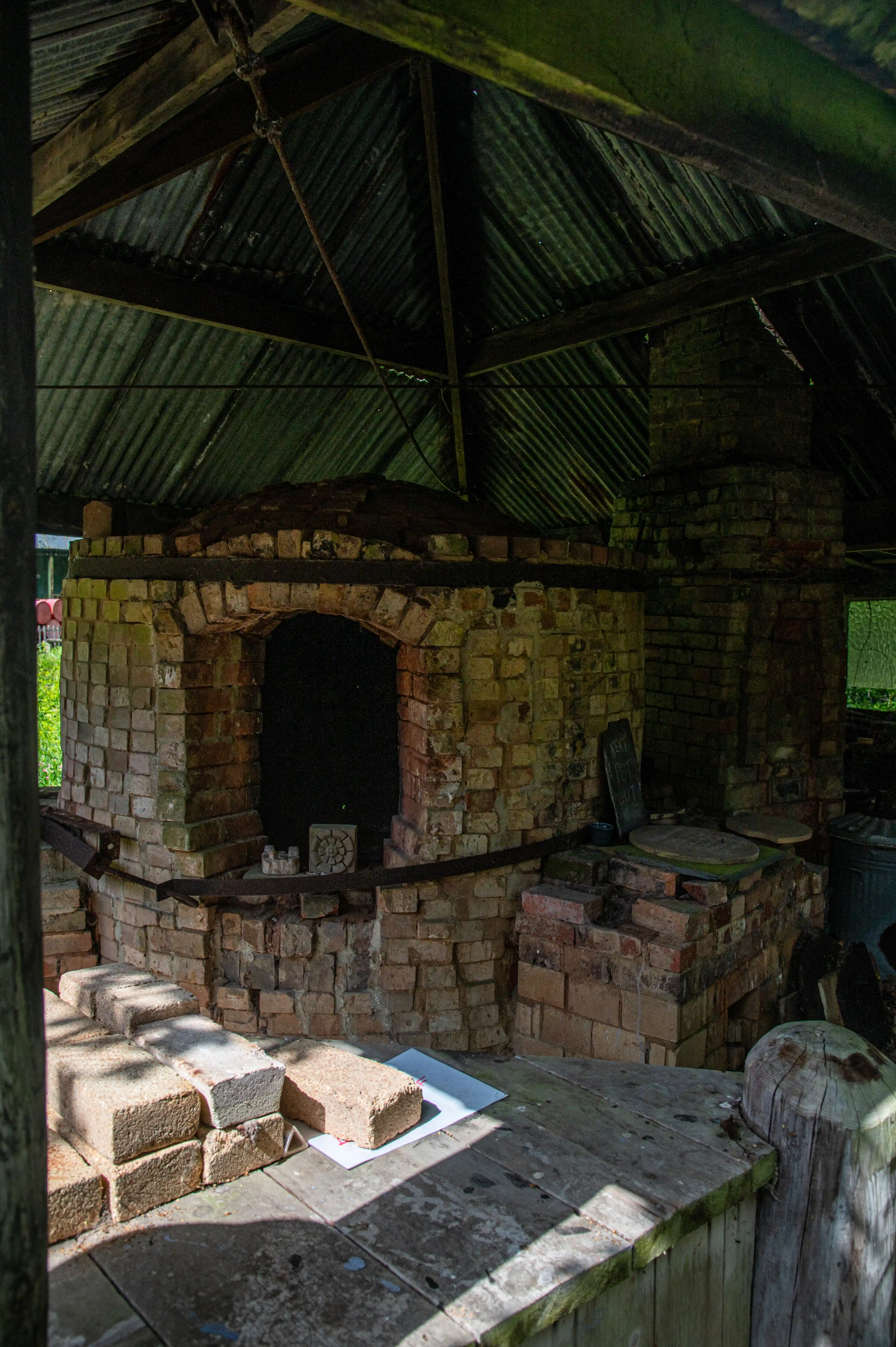 A rustic brick kiln with a large arched entrance and a chimney, covered by a corrugated iron roof. A wooden table in front of the kiln holds a stack of bricks and a sheet of paper.