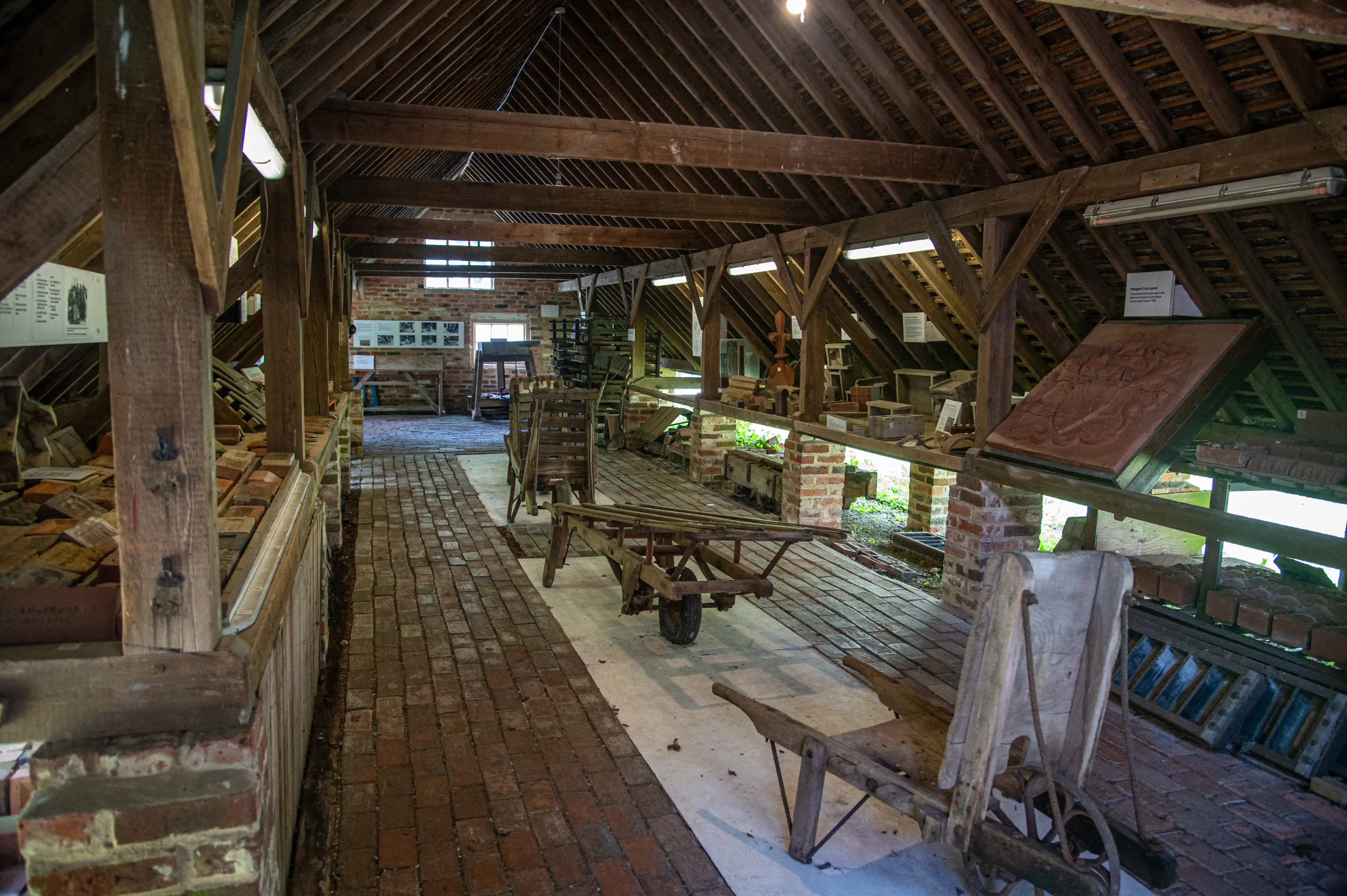 Interior of an old wooden building with exposed timber beams, brick walls, and brick floor. Vintage industrial equipment such as wooden wheelbarrows and displays of bricks are visible.
