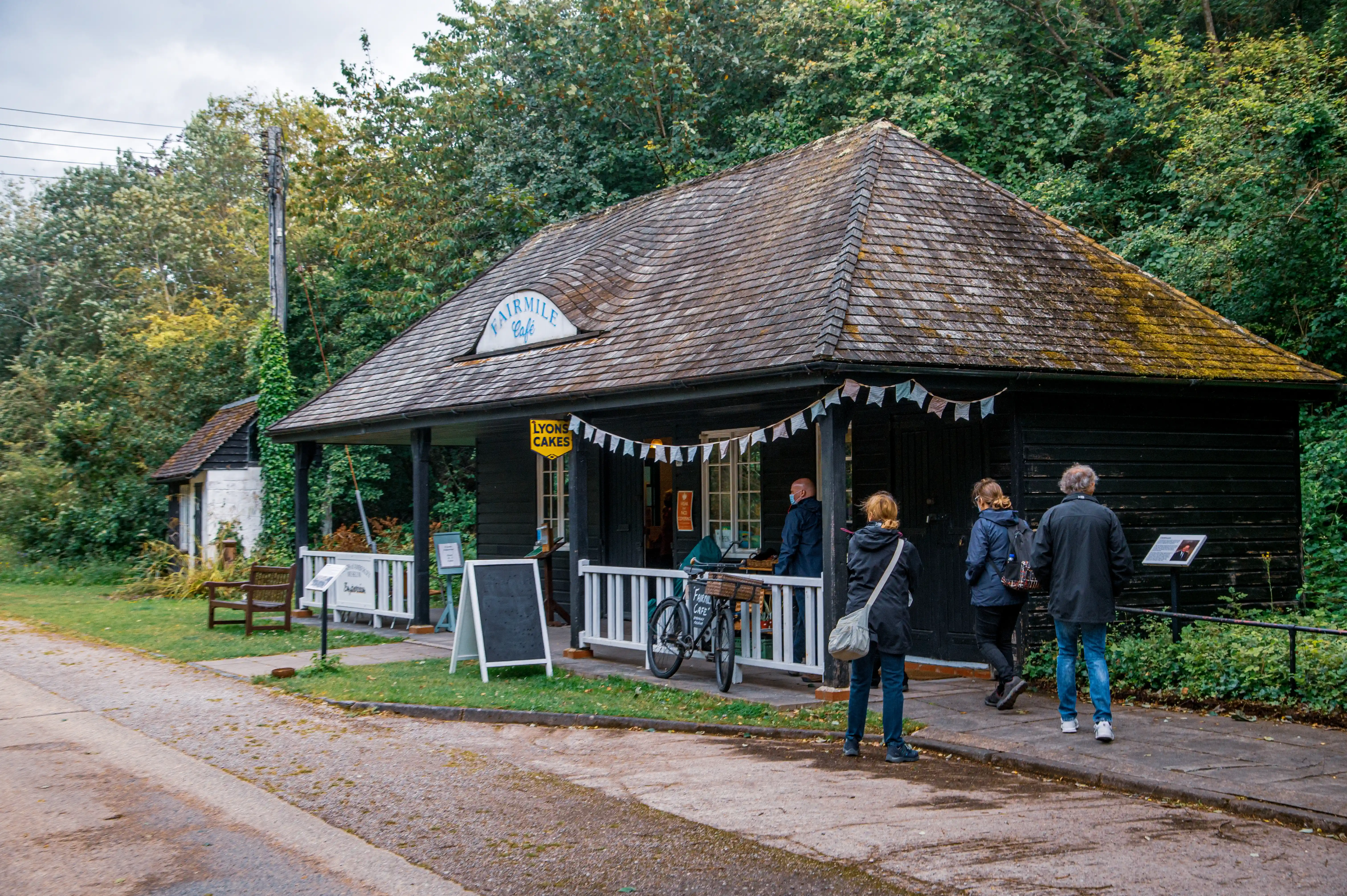 The exterior of the Fairmile Cafe and Lyons Cakes shop, a dark wooden building with a shingled roof, on a cloudy day. Several people are walking towards the entrance, and a bicycle is parked on the porch.