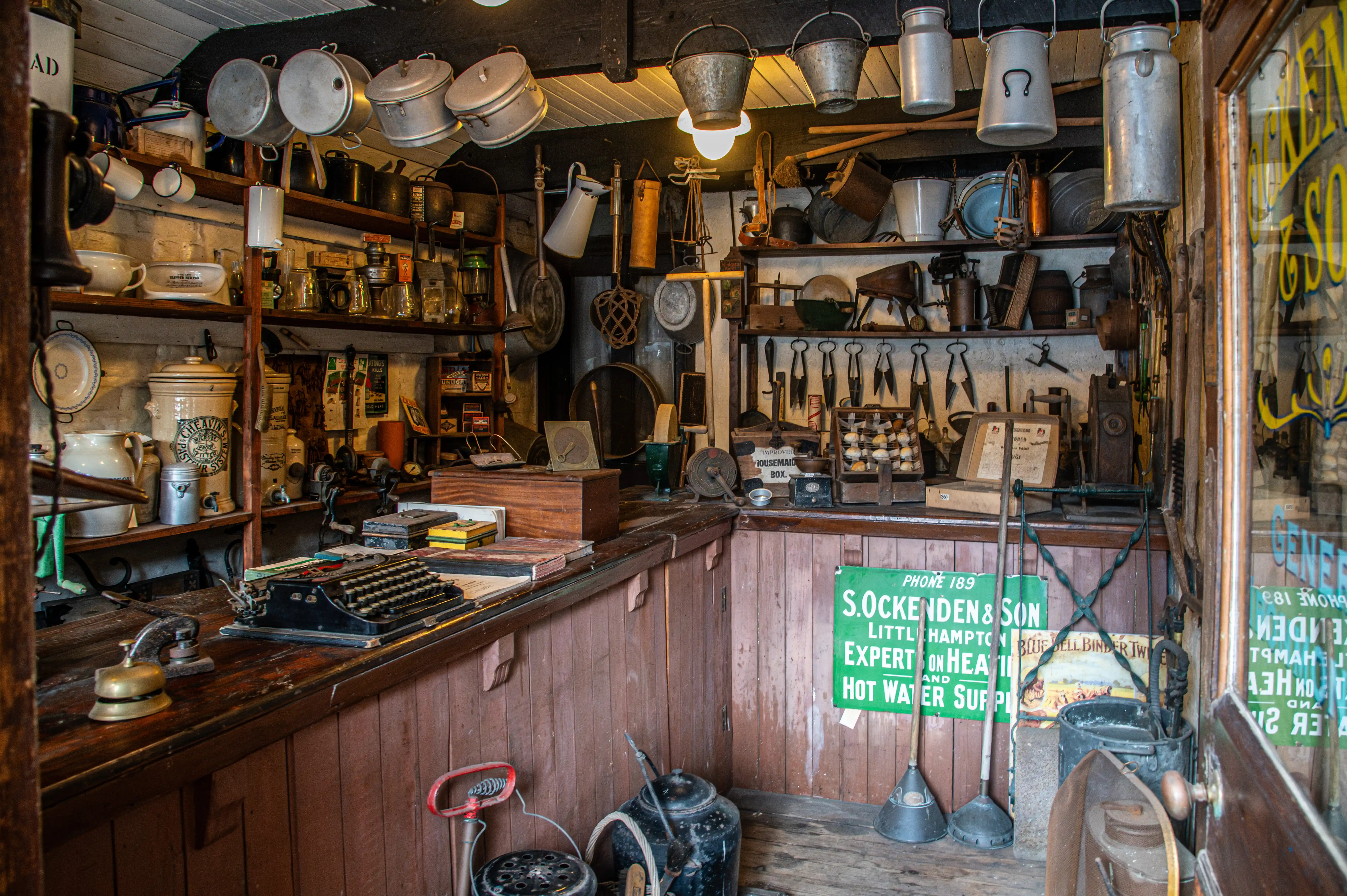 Interior view of a cluttered vintage general store with shelves filled with pots, pans, tools, and other historical items. A vintage typewriter sits on the counter.