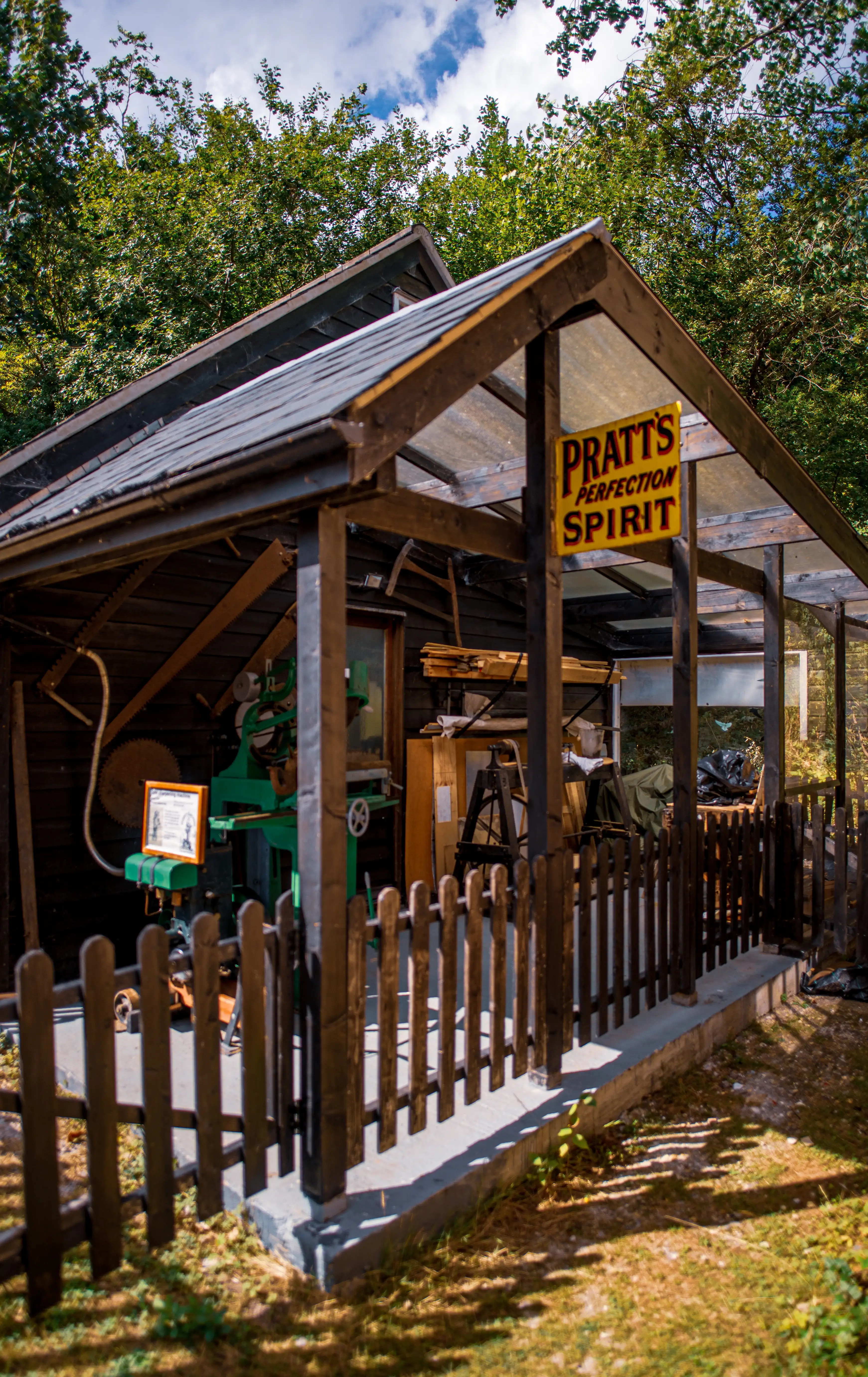 Exterior view of an old, dark wooden workshop with a partially open roof structure. A yellow sign reading 