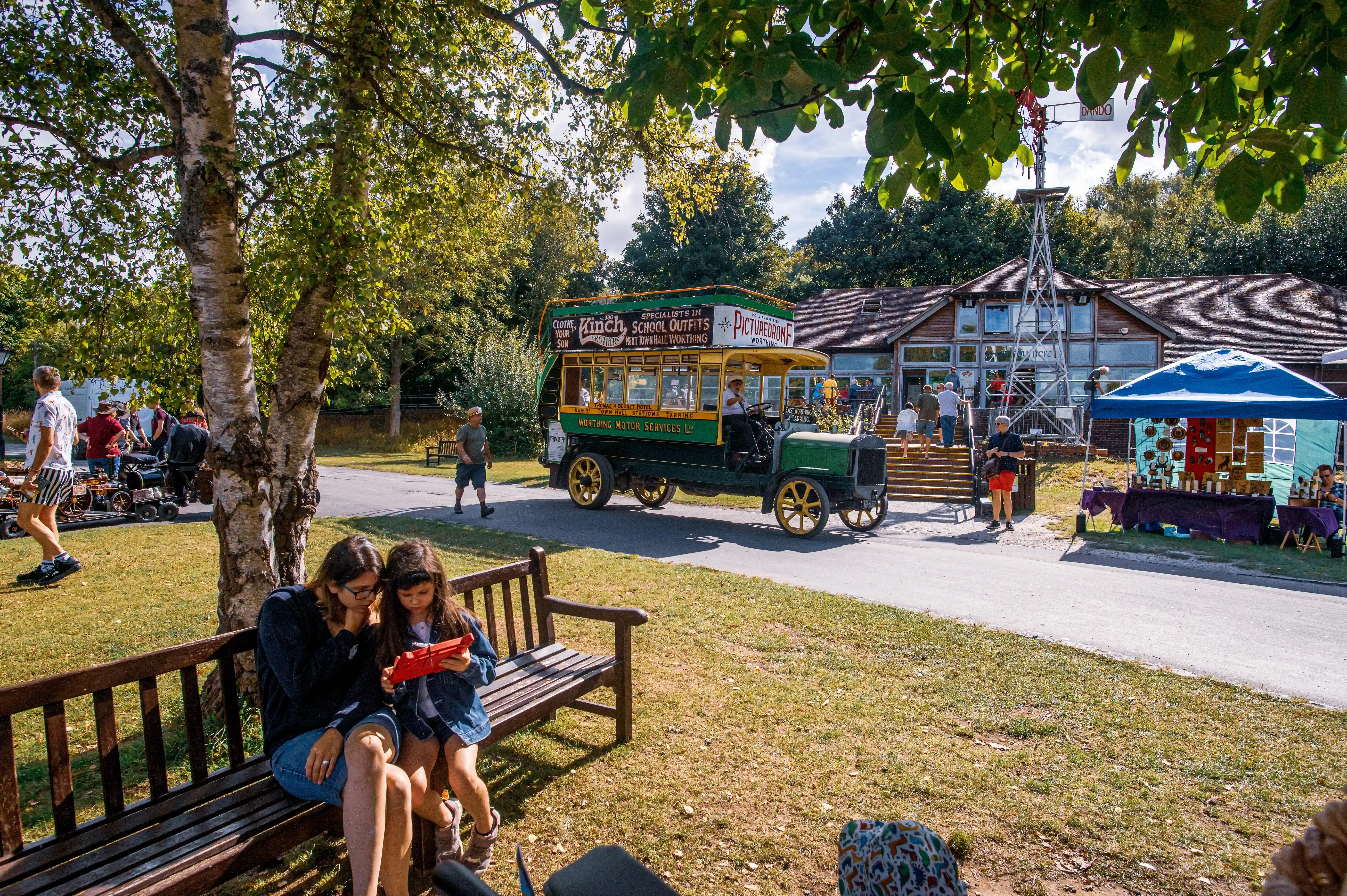 Two girls sit on a park bench looking at a red object, with a vintage green and yellow double-decker bus and a gathering of people in the background.