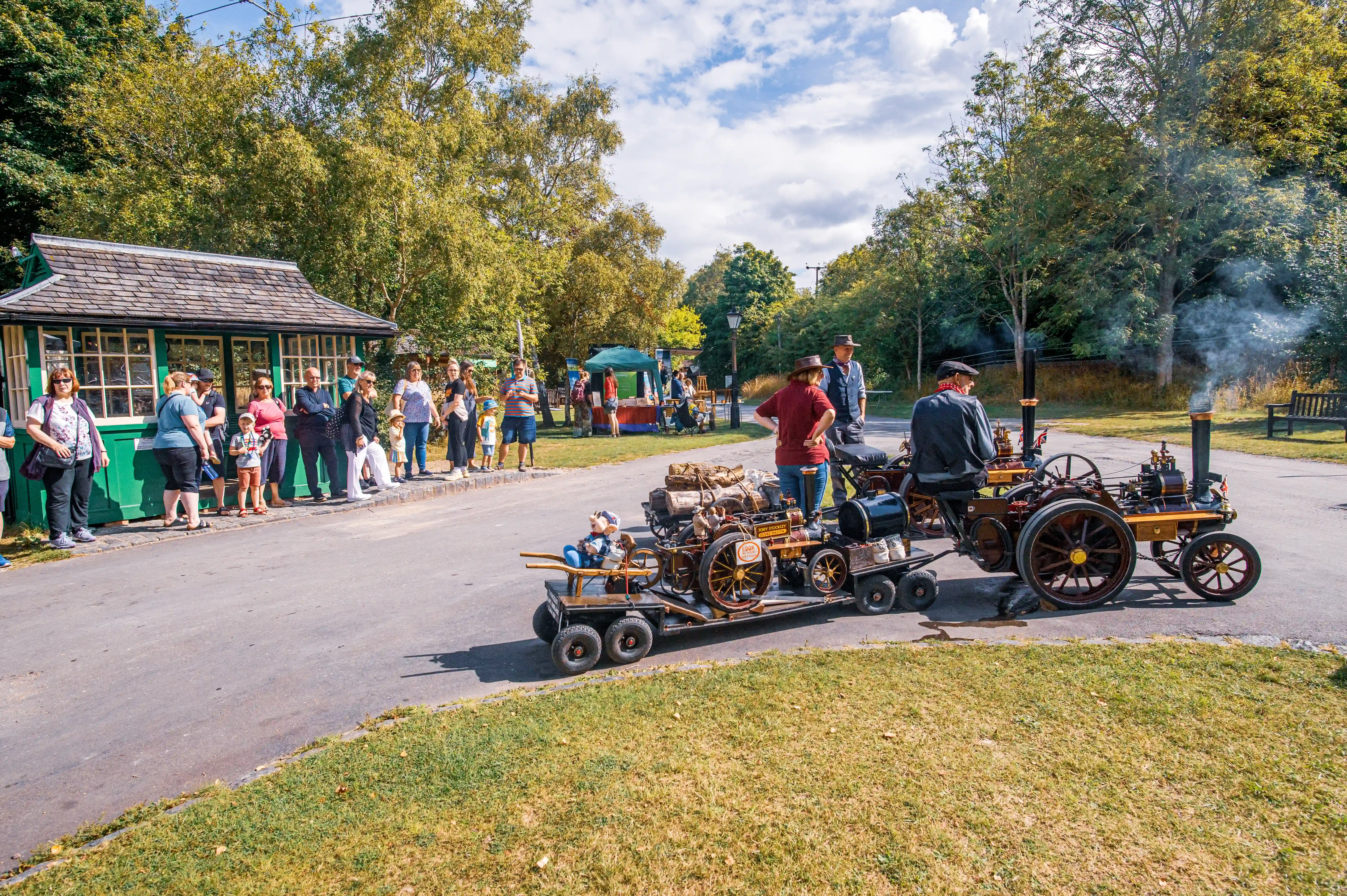 A sunny outdoor event with a crowd of people watching two vintage steam traction engines, one pulling a trailer with a stuffed figure.
