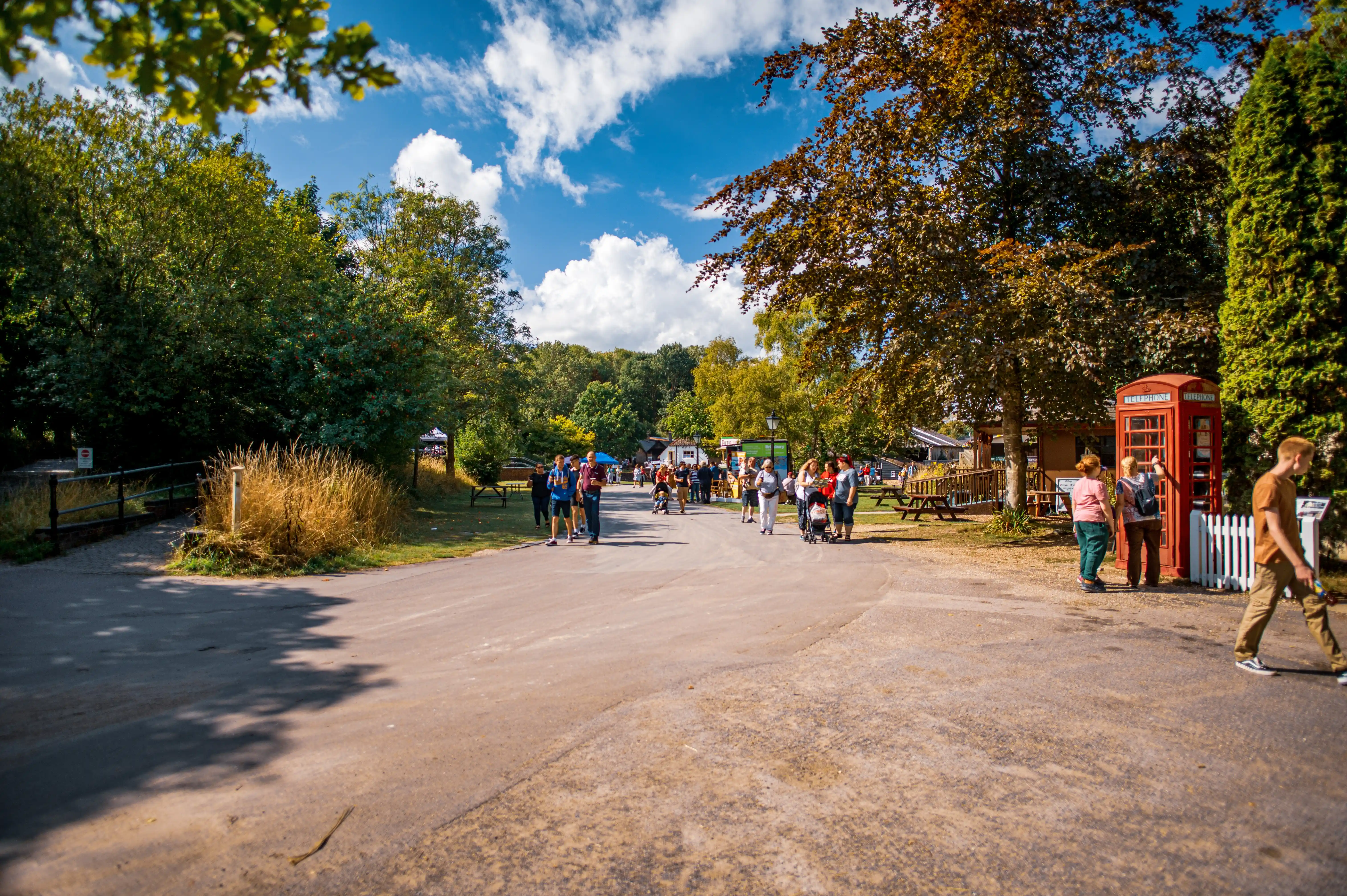 A wide shot of an outdoor recreational area on a sunny day, with a road leading into the distance where a group of people are walking. To the right, a bright red British telephone box stands next to a white picket fence. Trees and foliage frame the scene.