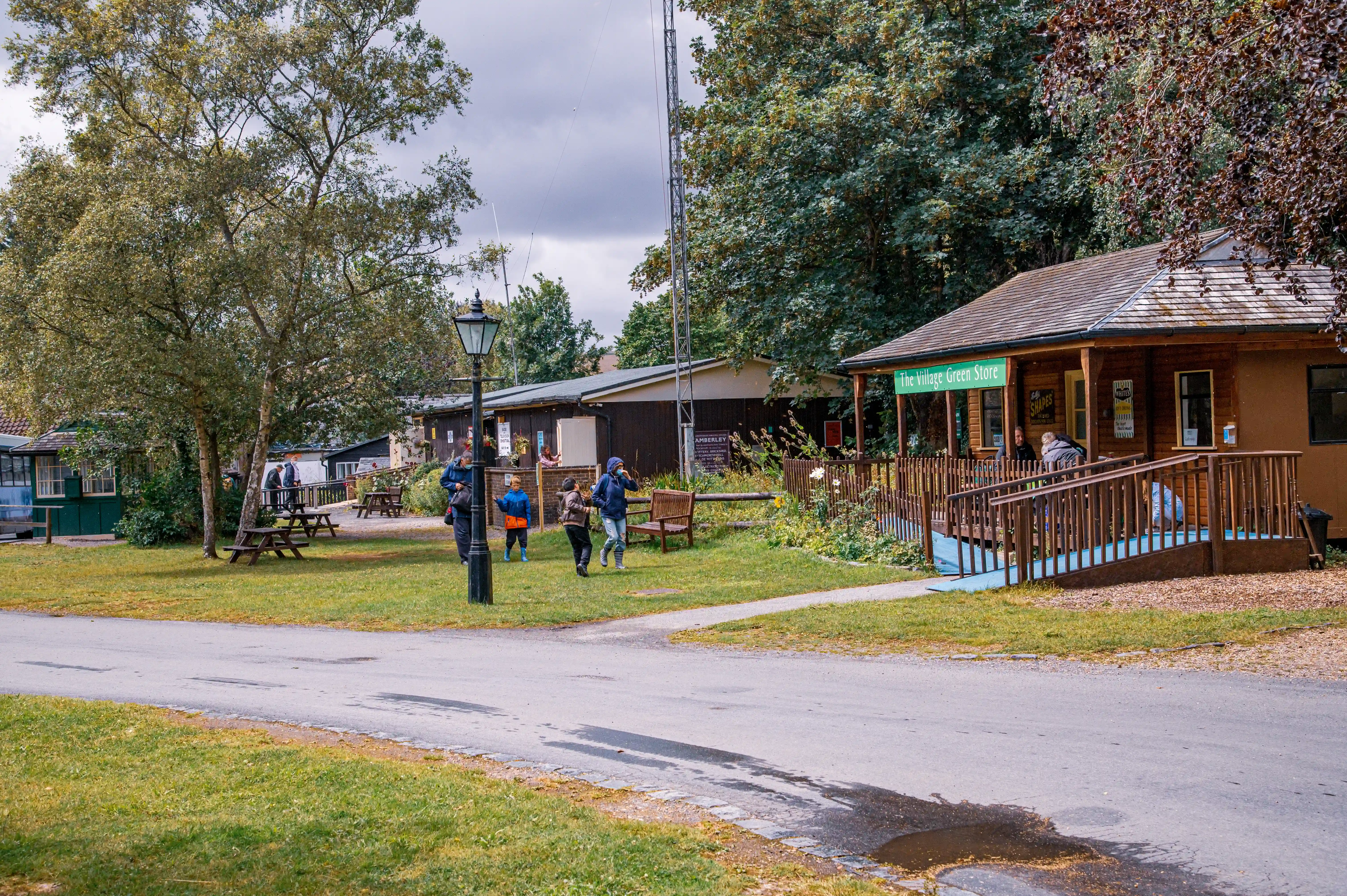 A wooden building with a sign reading 'The Village Green Store' in a lush, green countryside. The store has a small porch with wooden railings and a ramp. Visitors, including children and adults, are walking and exploring the area, which includes picnic tables, a lamppost, and trees