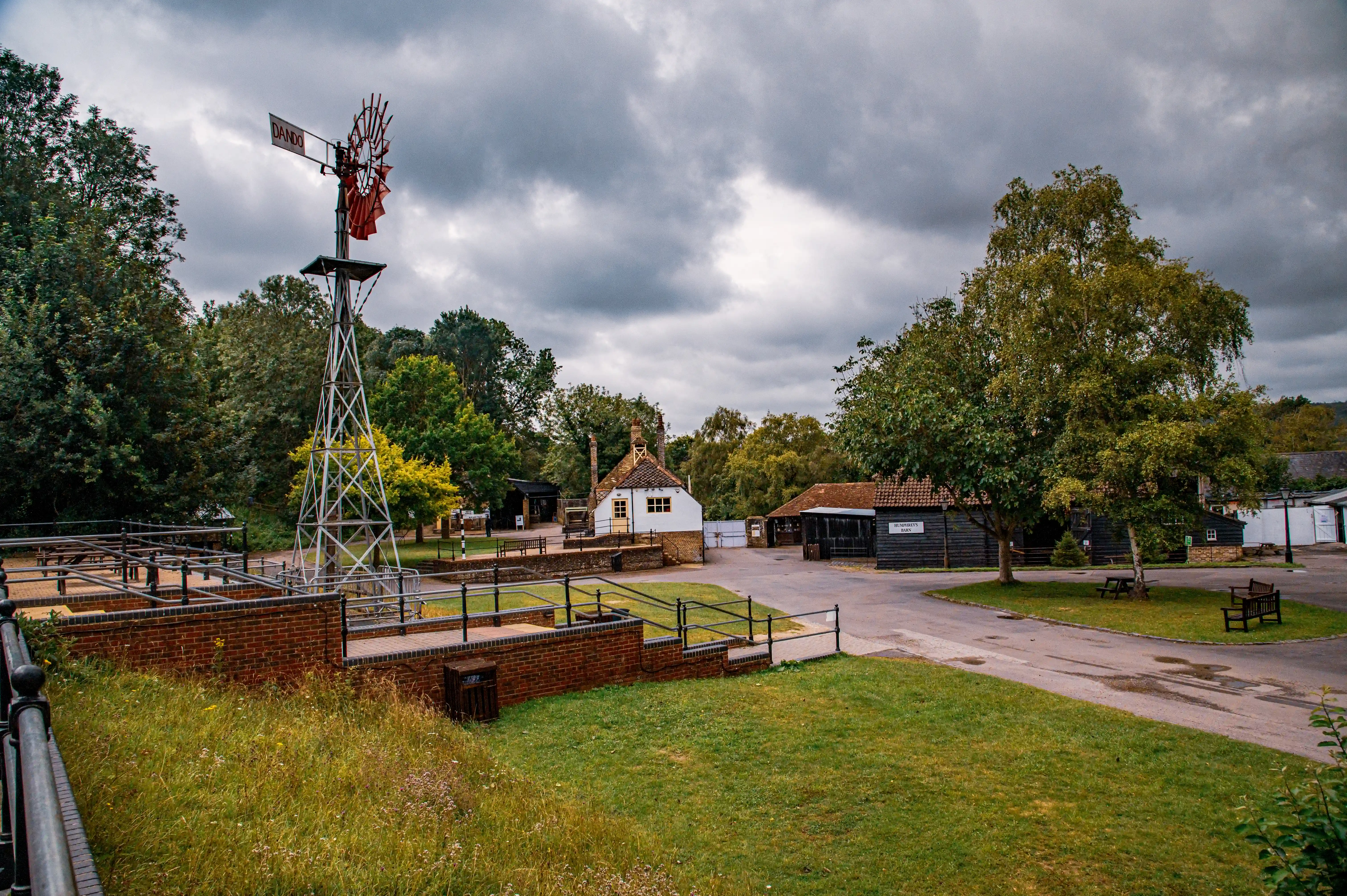 A vintage metal windmill with red sails is situated in the foreground, overlooking a courtyard with several traditional buildings, including a white cottage with orange-framed windows and dark wooden barns. The scene is set against a backdrop of lush green trees and a stormy grey sky.