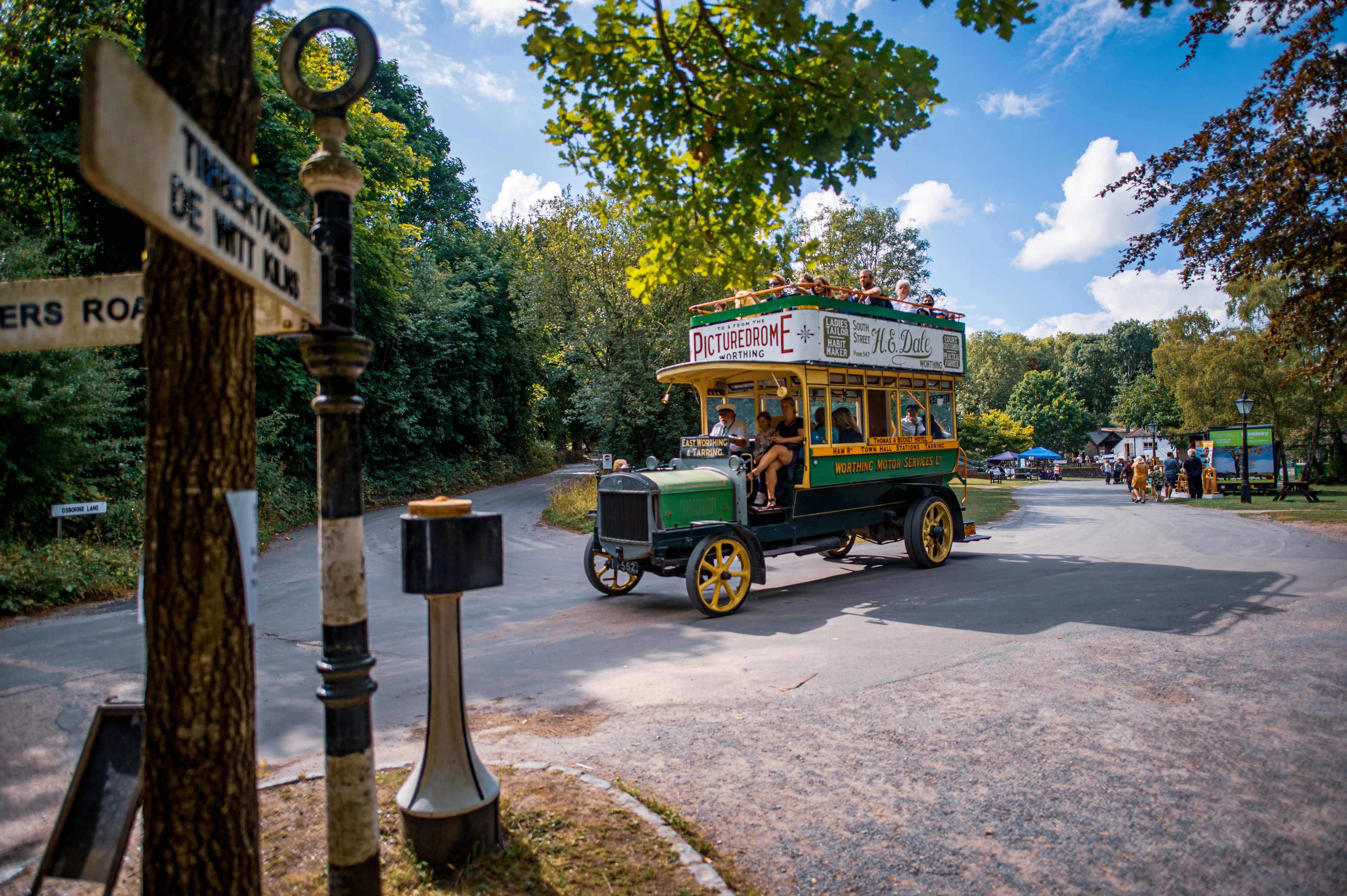 A vintage green and yellow open-top bus, carrying passengers, drives down a paved road. To the left, a wooden signpost with directional arrows is attached to a tree. The background features dense green foliage and a bright blue sky with white clouds.