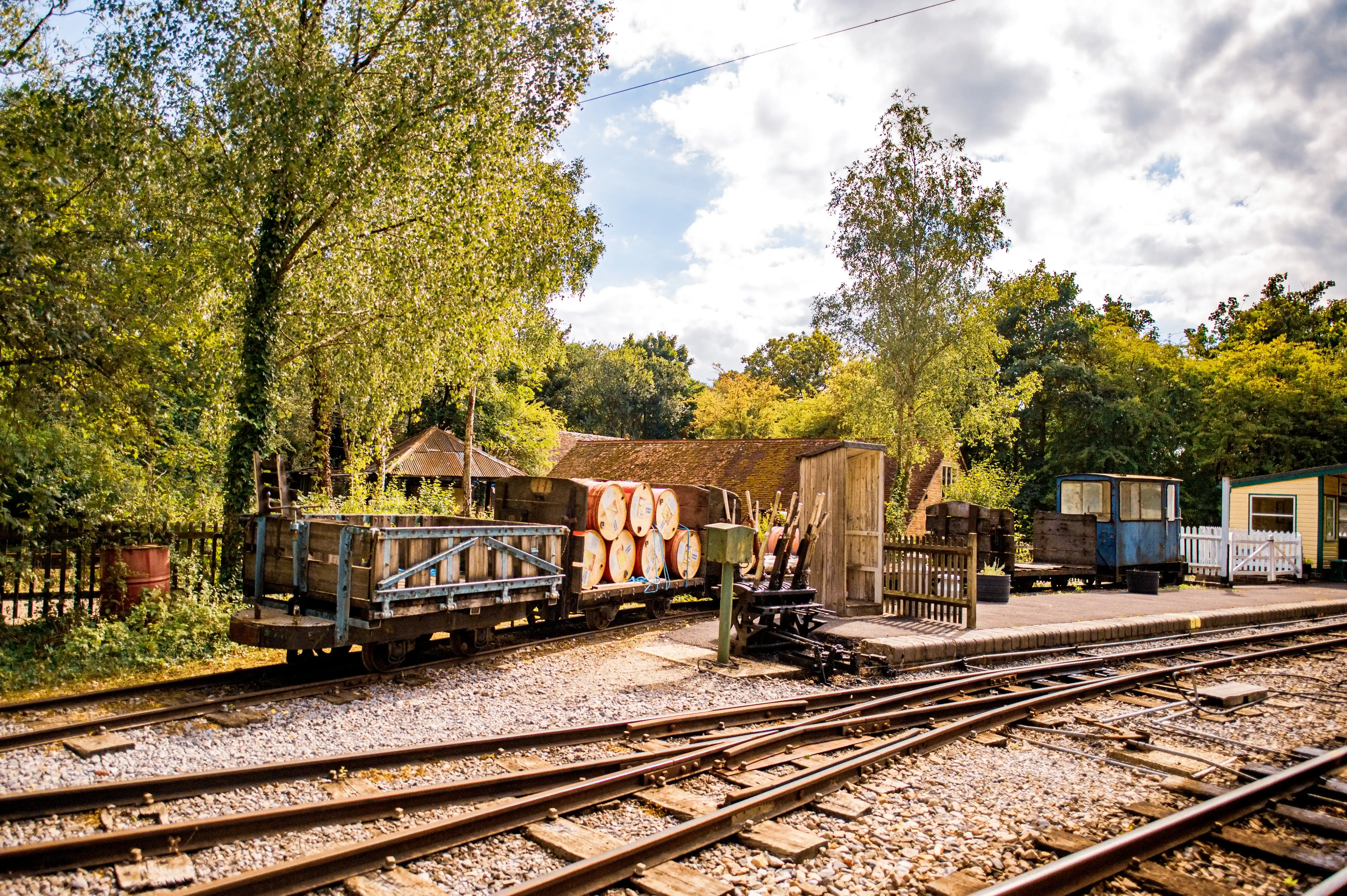 A railway wagon filled with orange barrels is parked on train tracks at a station. A blue carriage and a yellow building are visible further down the platform, surrounded by trees and under a cloudy sky.