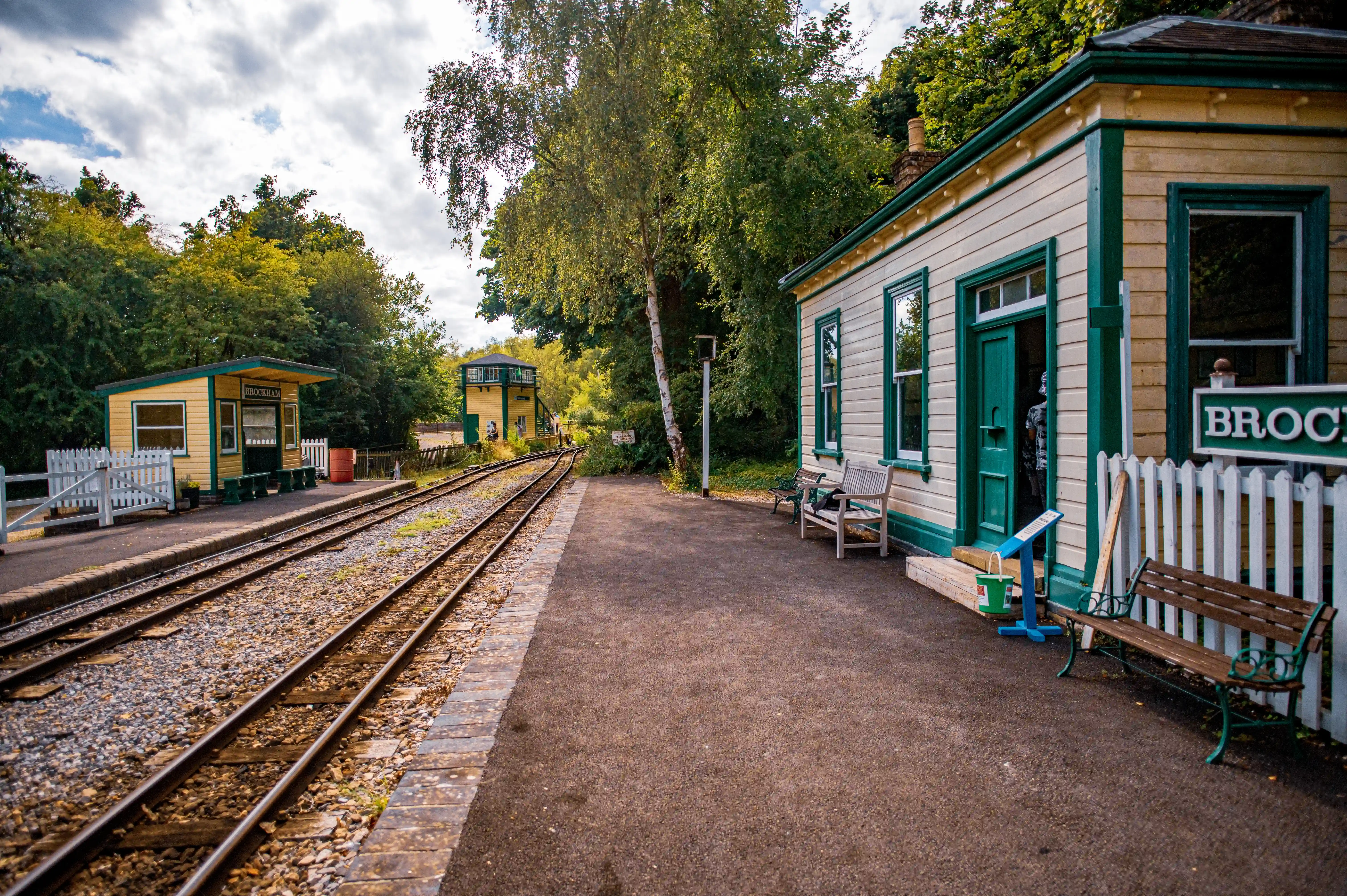 A vintage railway station with a pale yellow building with green trim, a covered platform, and train tracks.