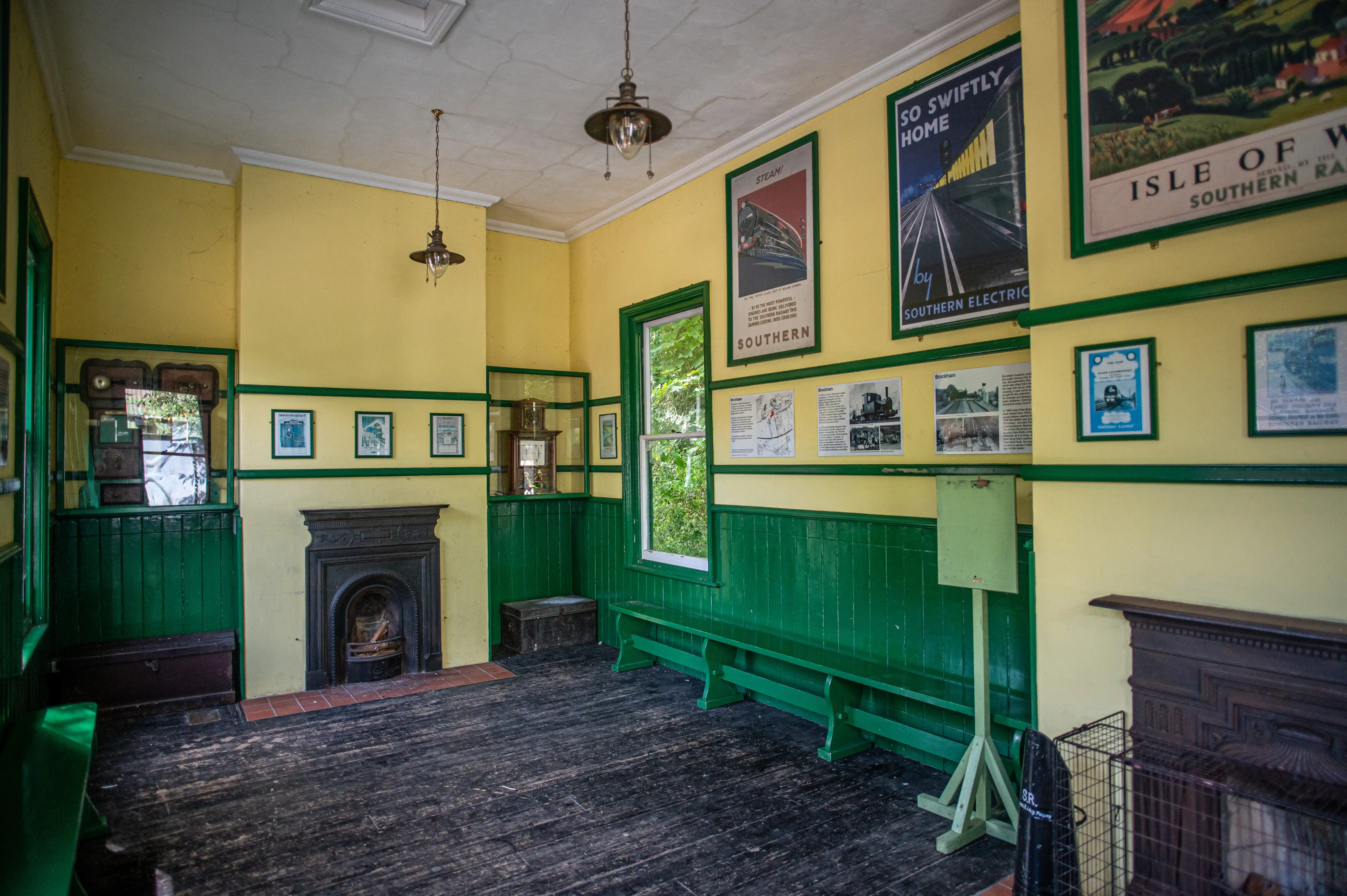 Interior of a historic railway station waiting room, with yellow walls, green wainscoting, and dark wooden floorboards. Vintage posters depicting train travel are displayed on the walls, alongside a fireplace and wooden benches.