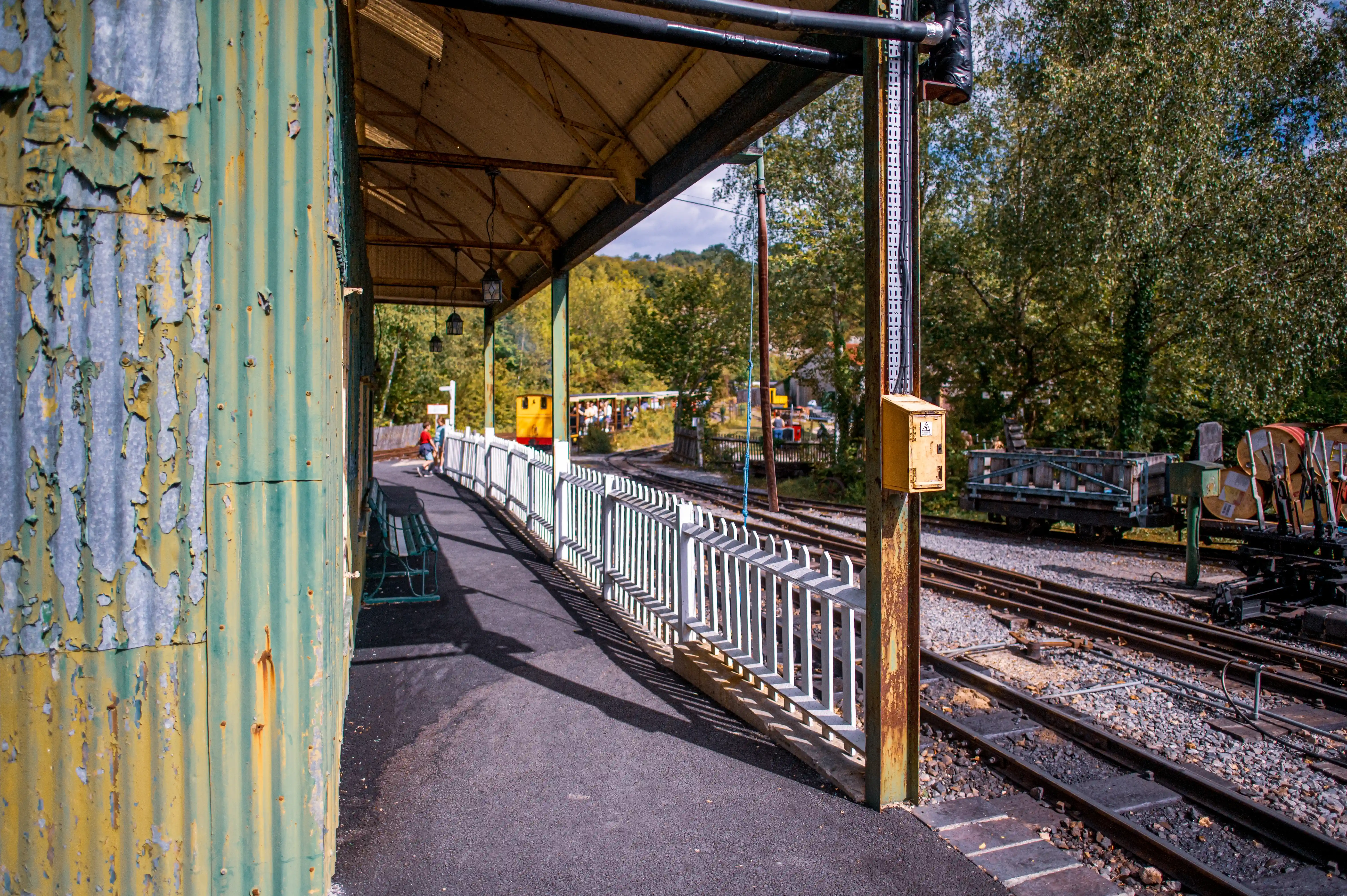 A view of a train station platform with a weathered corrugated iron wall on the left, a white railing, and train tracks with a yellow train in the background.
