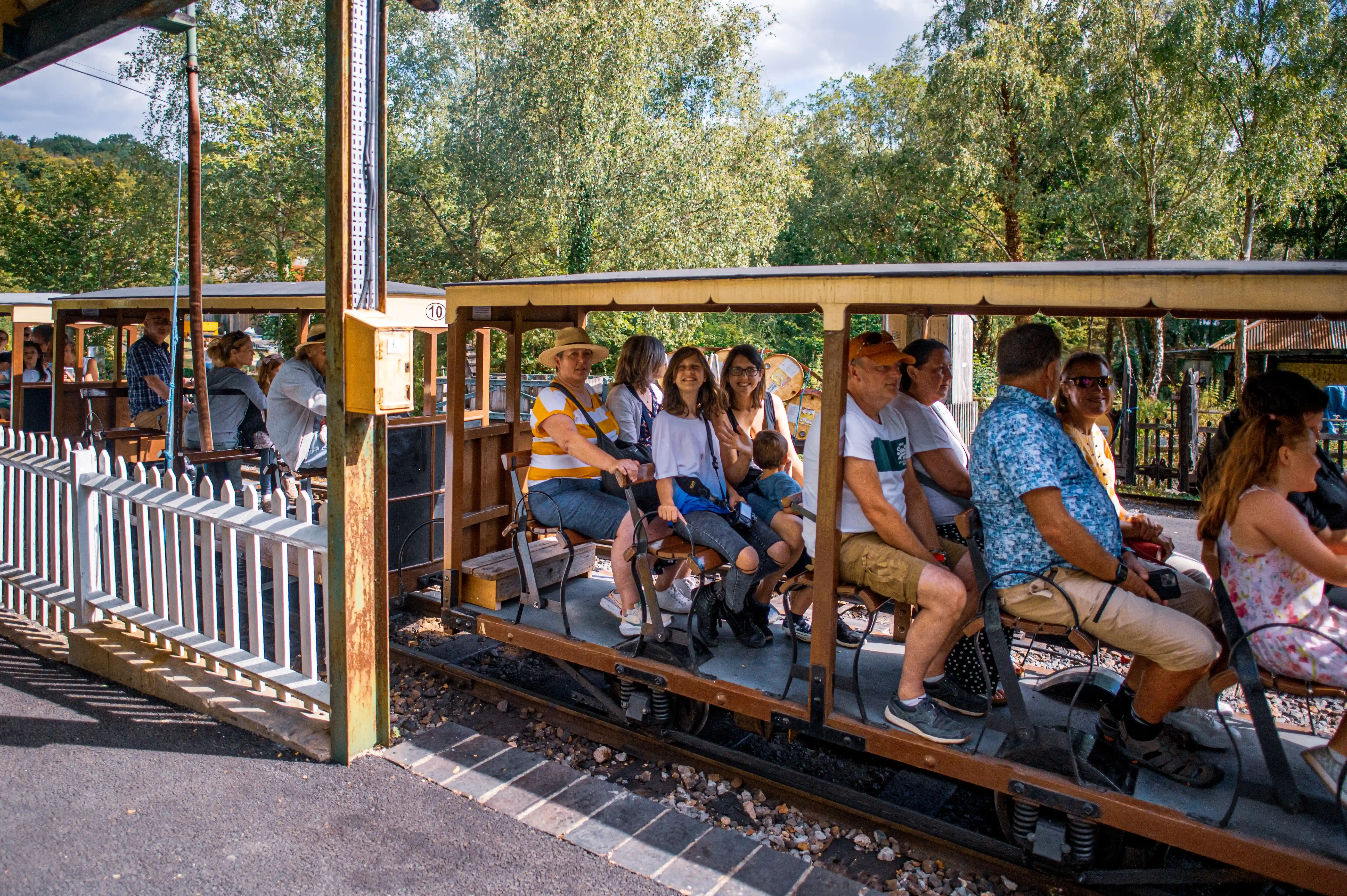 A group of passengers, including adults and children, riding in an open-air heritage railway carriage. The carriage has wooden seats and metal railings, and the passengers are dressed casually, some wearing hats and carrying bags. The train travels through a lush, green landscape.