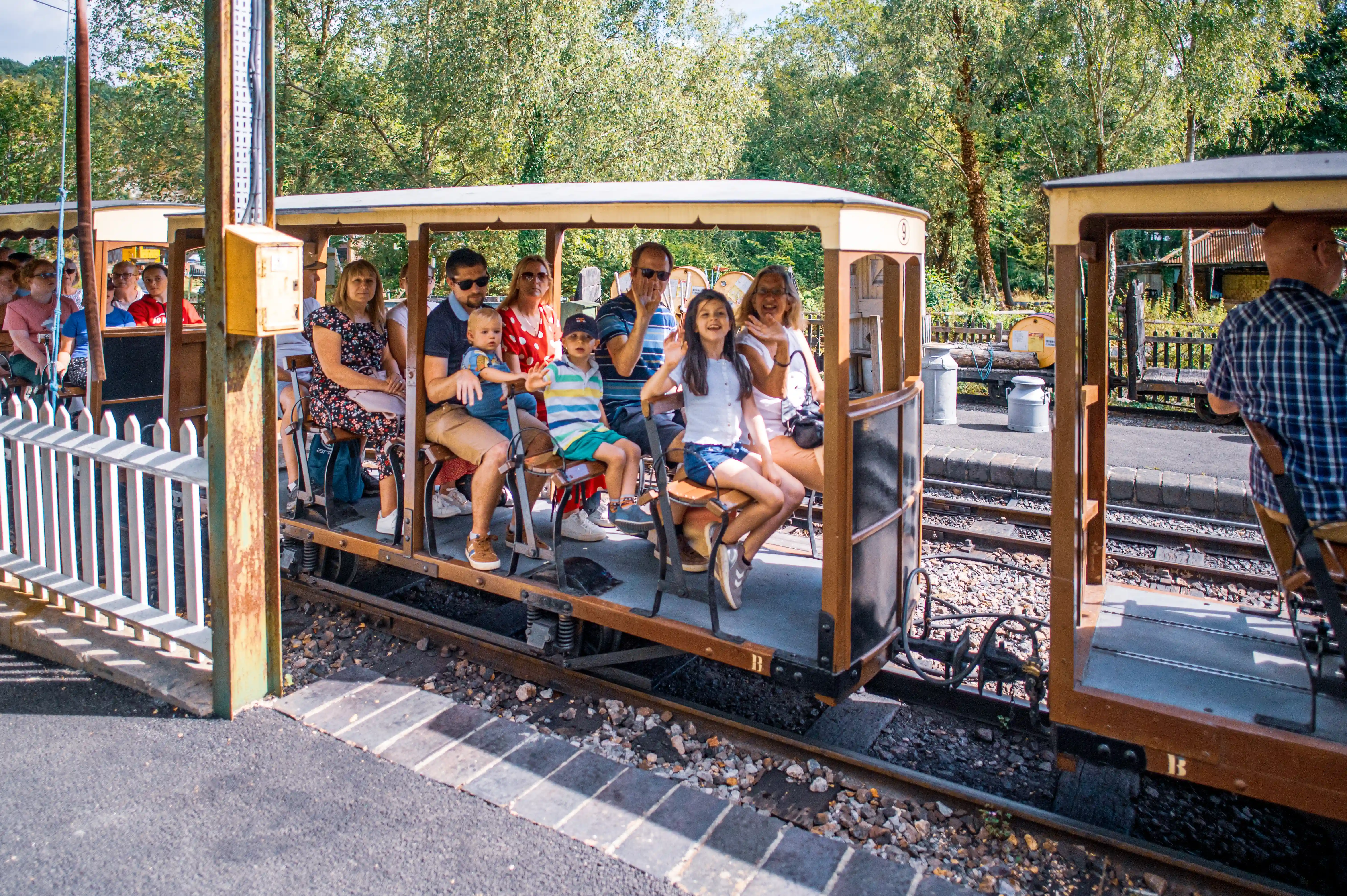 A diverse group of people, including adults and children, are seated together on a vintage miniature train carriage with an open-air design. They are on tracks surrounded by greenery, and several individuals are smiling and waving.