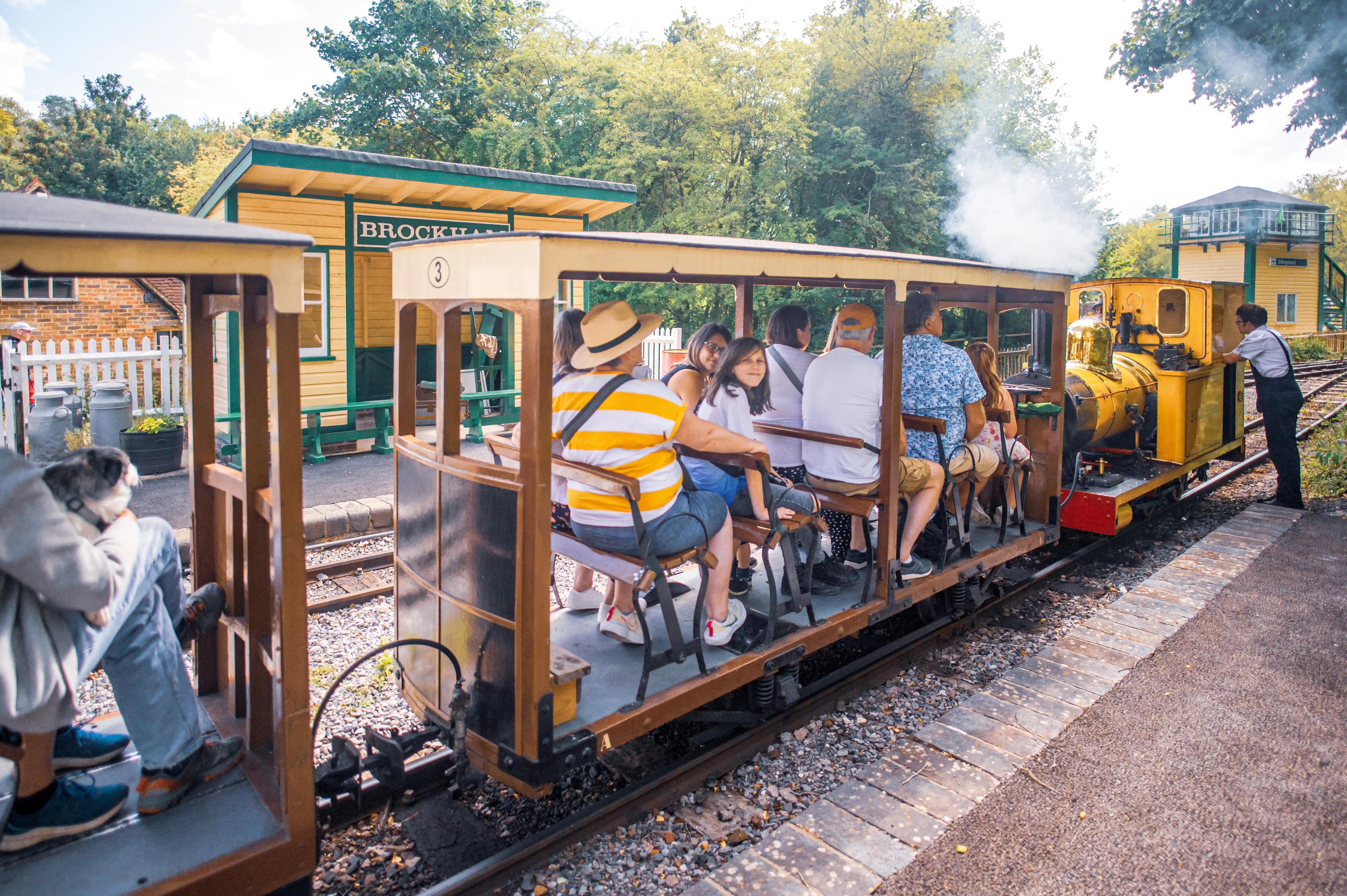 A miniature steam train with several passengers in an open carriage travels along a track past Brockham station.