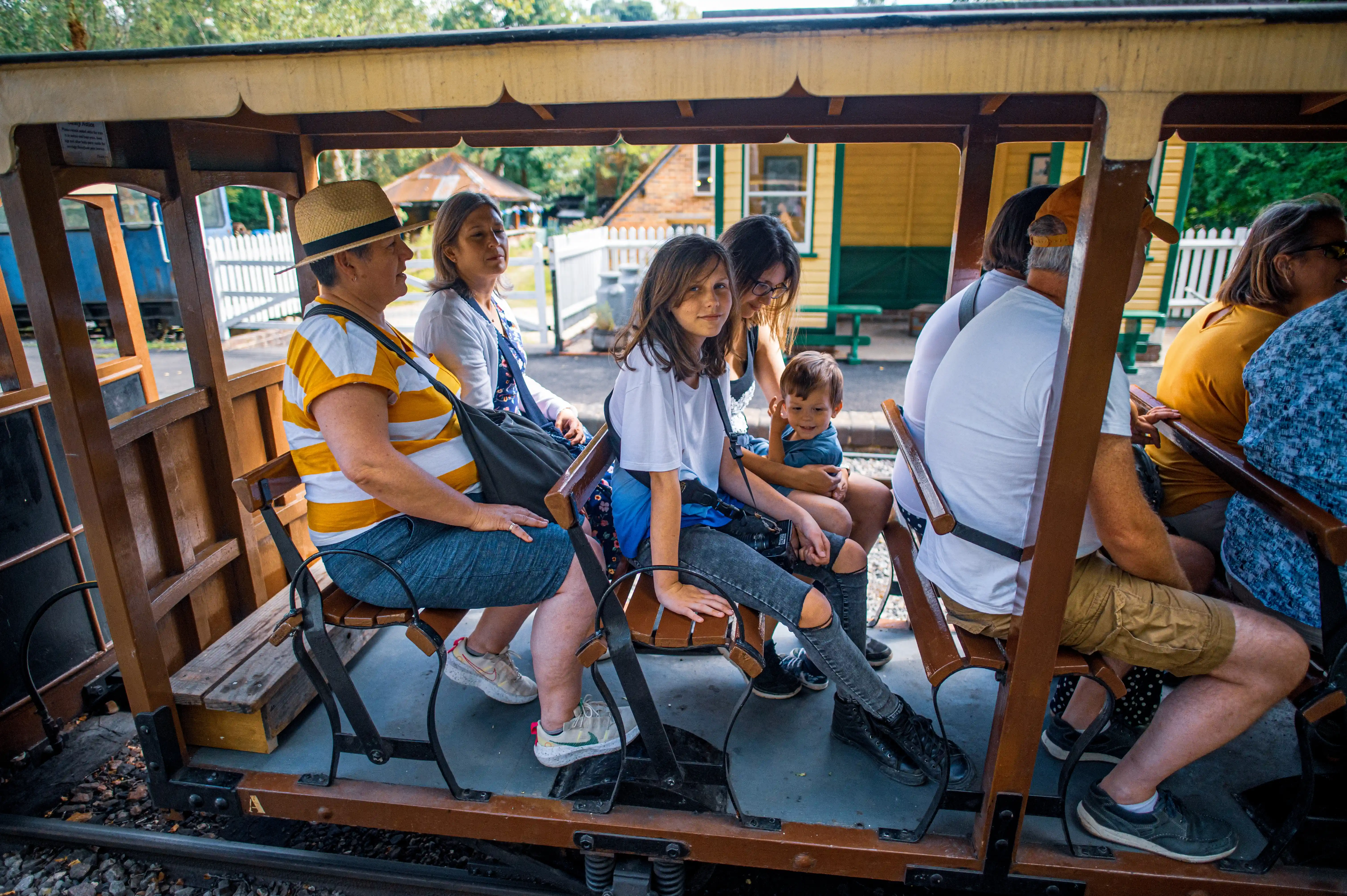 A group of people, including men, women, and children, are seated on an open-air vintage train carriage. They are wearing casual clothing and appear to be enjoying a journey.