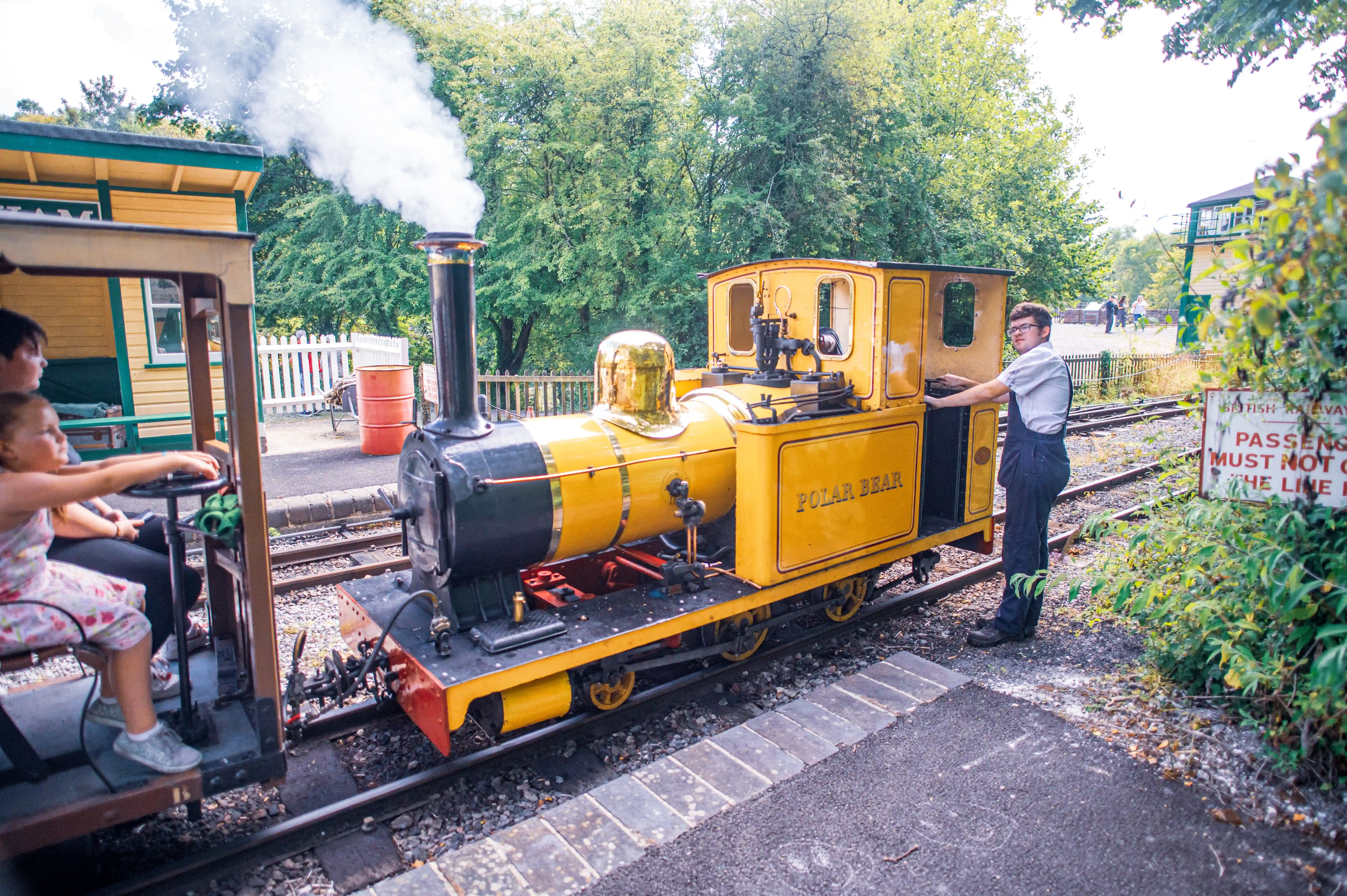 A bright yellow miniature steam train labelled 'Polar Bear' is stopped at a station. A young girl is seated in the driver's cab, holding onto the steering wheel. A young man in dark overalls and a light shirt stands next to the train, his hand on the open door. Steam is rising from the train's chimney, and a railway signal box and trees are visible in the background.