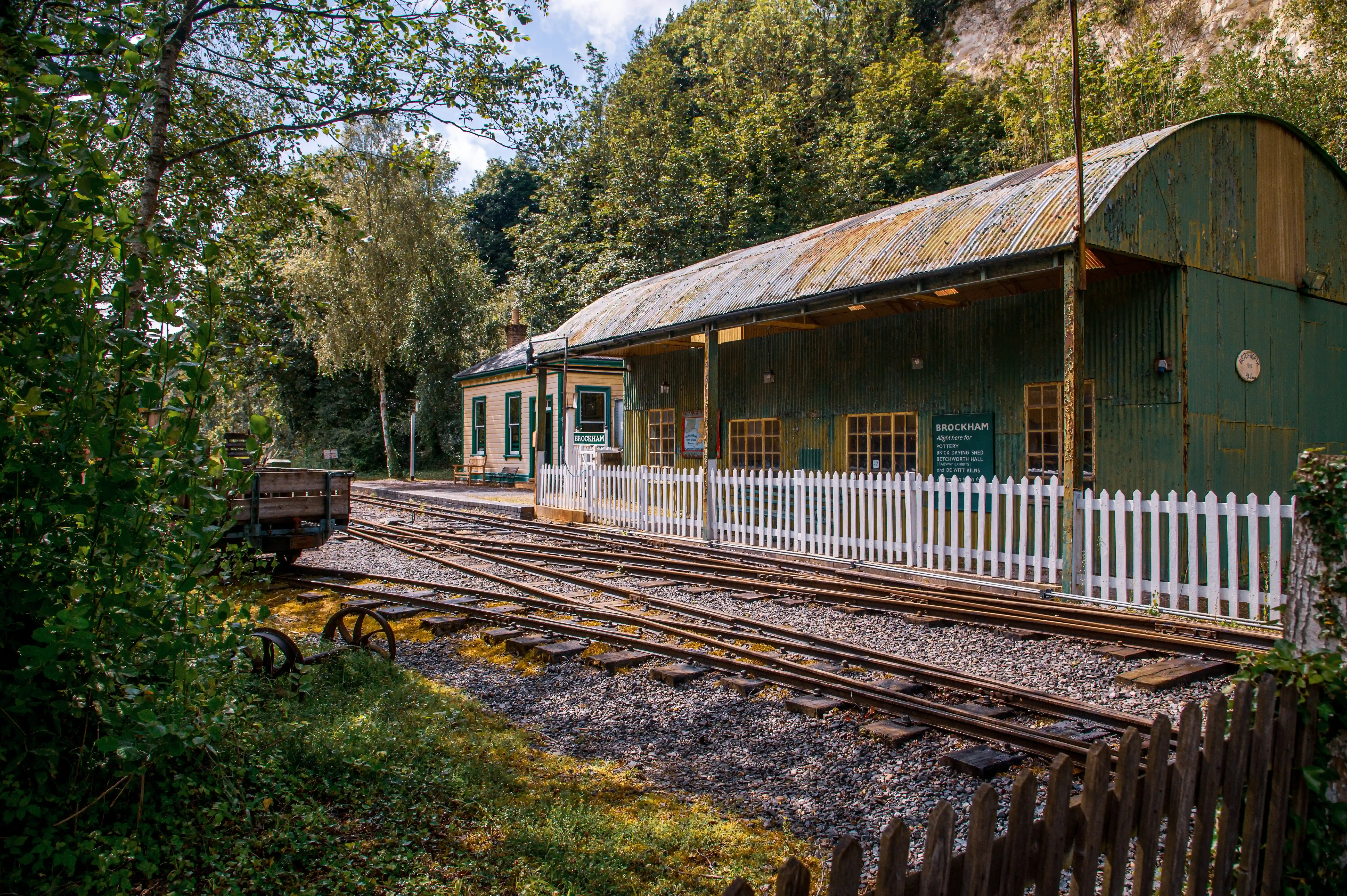 A wide shot of a miniature railway station with two sets of tracks leading towards a long, single-storey building with a green, corrugated iron roof. A white picket fence runs along the platform in front of the building. Trees and foliage surround the station, with a small, old-fashioned wagon visible on the left.