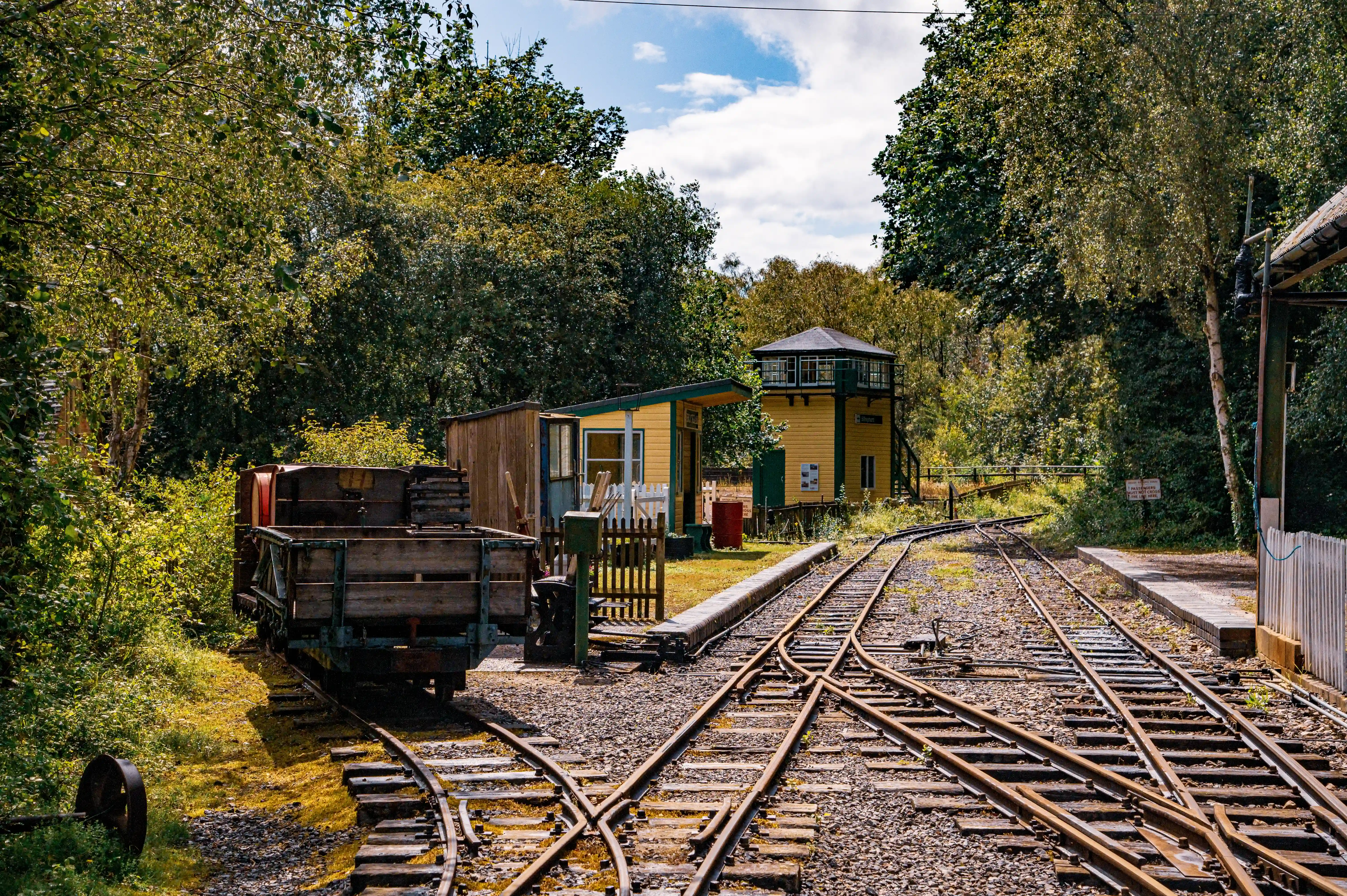 A view of a miniature railway station with multiple tracks diverging, a rustic wooden goods wagon, a small yellow building with a tower, and surrounding trees.