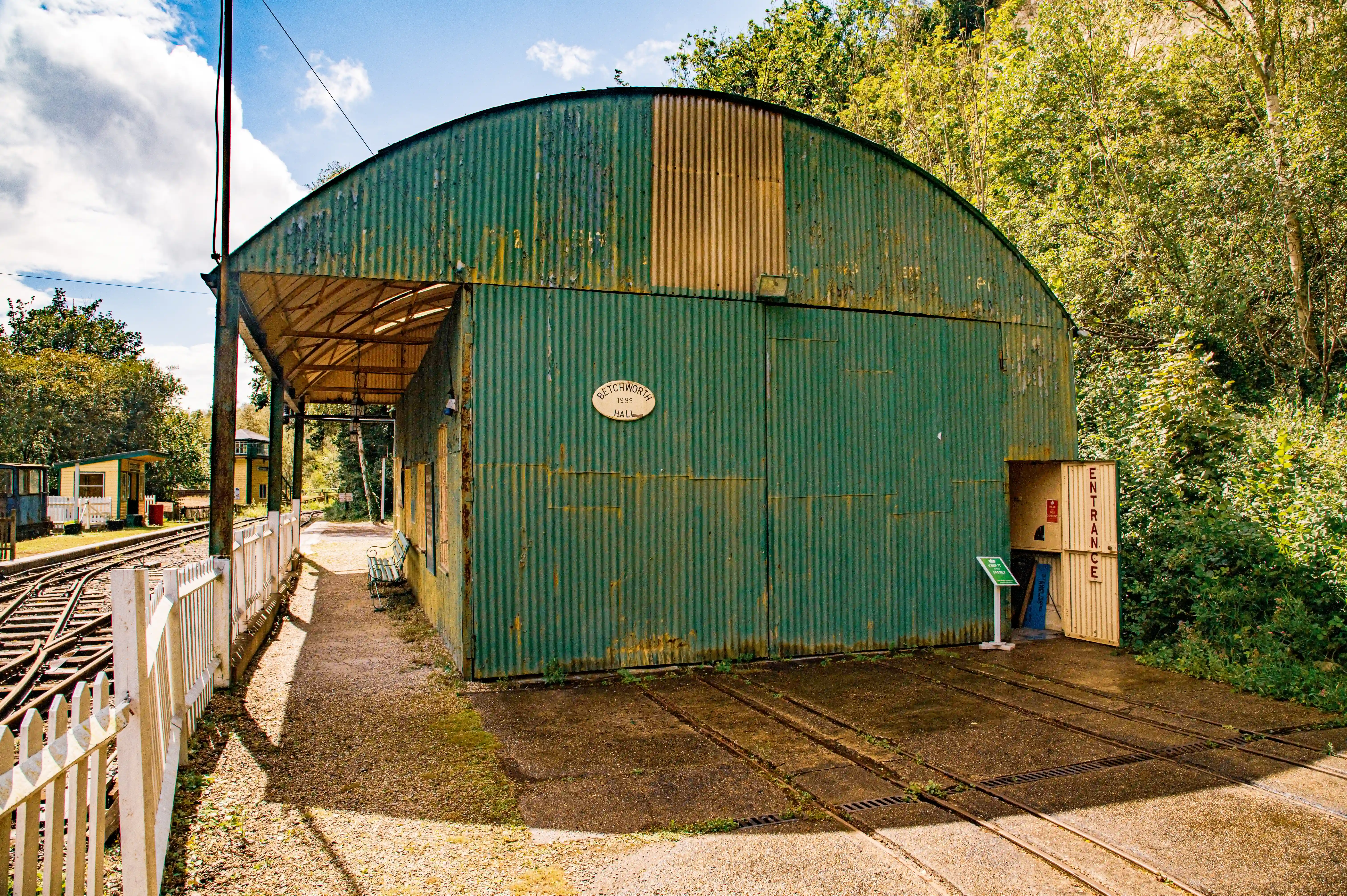 A green, arched corrugated iron railway shed with yellow accents sits next to a train track and a white picket fence.