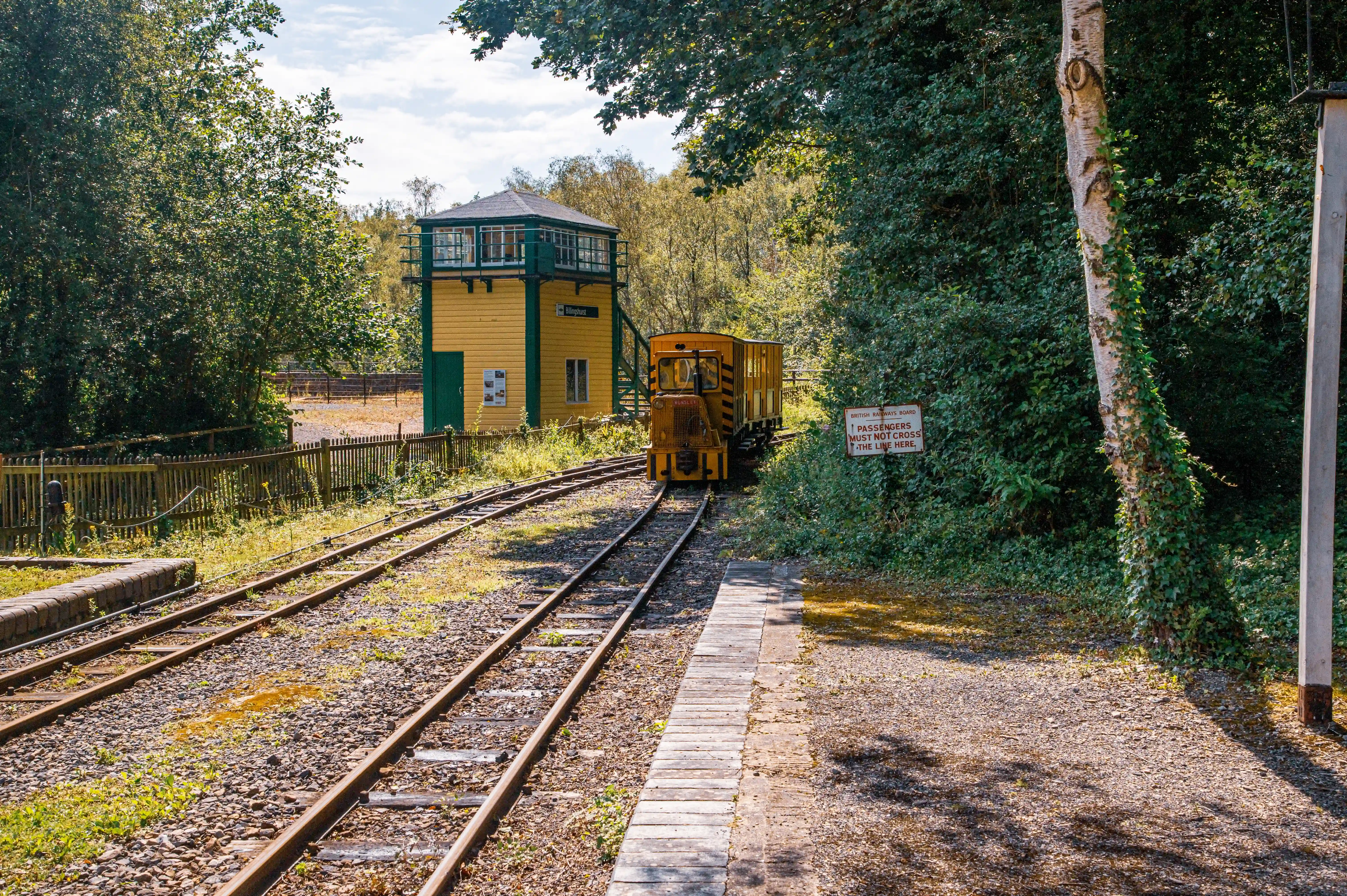 A narrow gauge train is approaching a signal box at a railway station. The train is yellow with an orange locomotive, and the signal box is painted yellow with green trim. There are trees and vegetation surrounding the station, and a platform is visible.