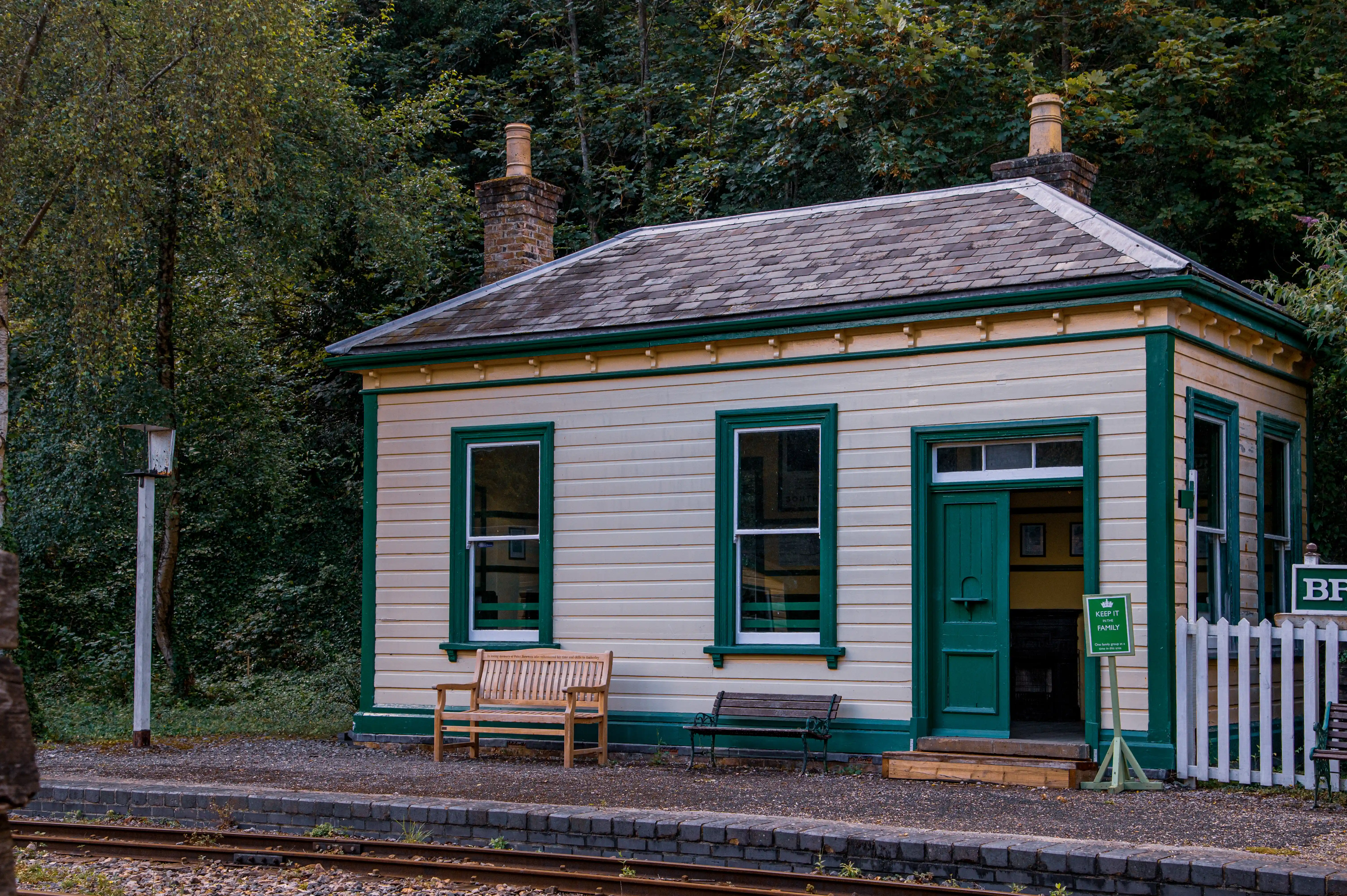 A small, vintage-style railway station building with cream siding and green trim, featuring two windows, an entrance, and a white picket fence, set against a natural background of trees.