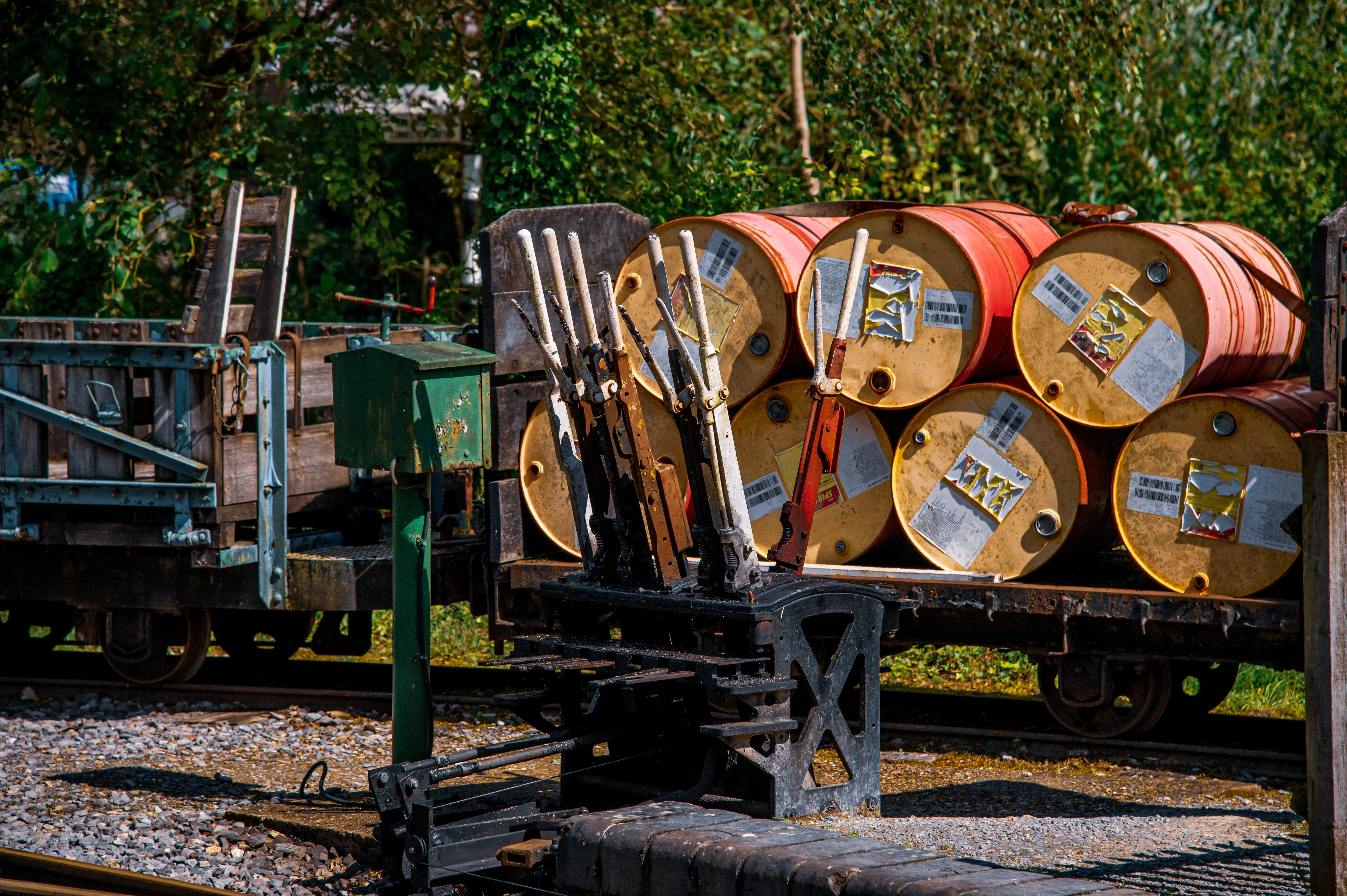 A close-up view shows several weathered railway signal levers mounted on a rusty metal platform. Behind the levers, a wagon is laden with a stack of large, round industrial barrels, some with labels. In the background, lush green foliage is visible.