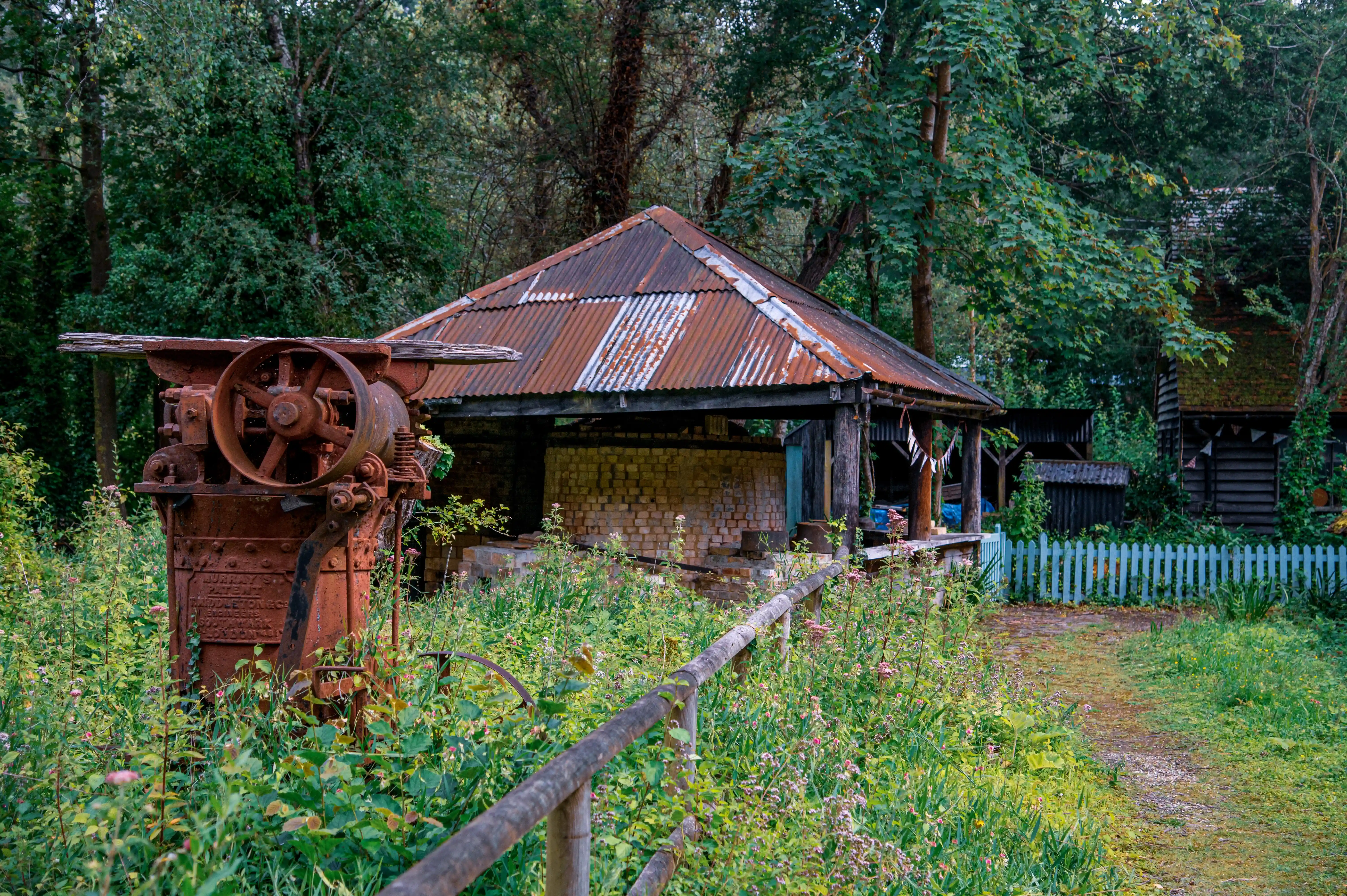 A close-up view of a weathered, rusty industrial machine with a large wheel, positioned in tall green grass and wildflowers. Behind it, an old brick structure with a corrugated iron roof and a rustic wooden building are visible amidst dense trees. A wooden fence runs along the right side of the frame.