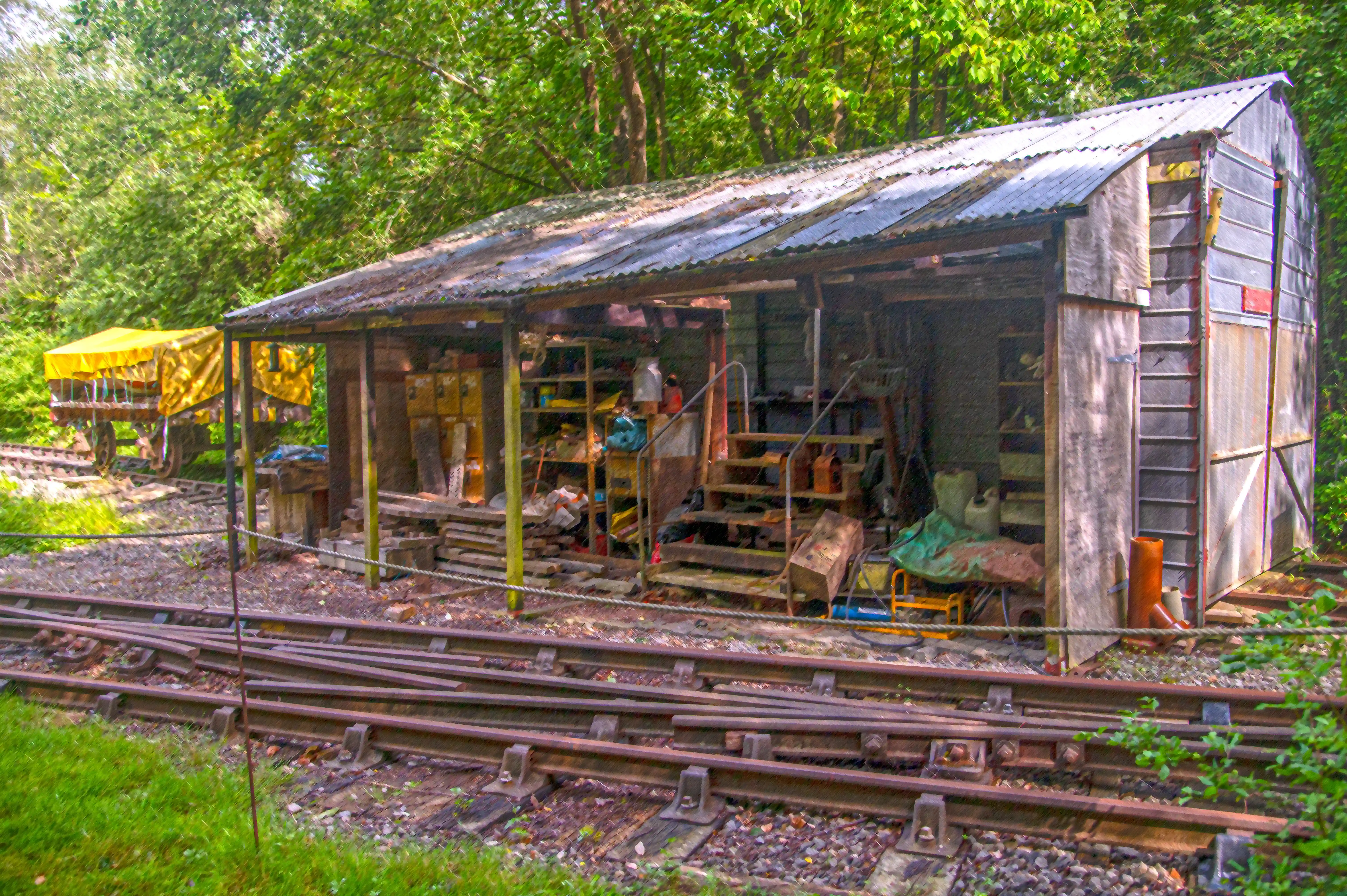 An open-fronted shed with a corrugated iron roof and wooden walls is filled with various items. Railway tracks run in front of the shed.