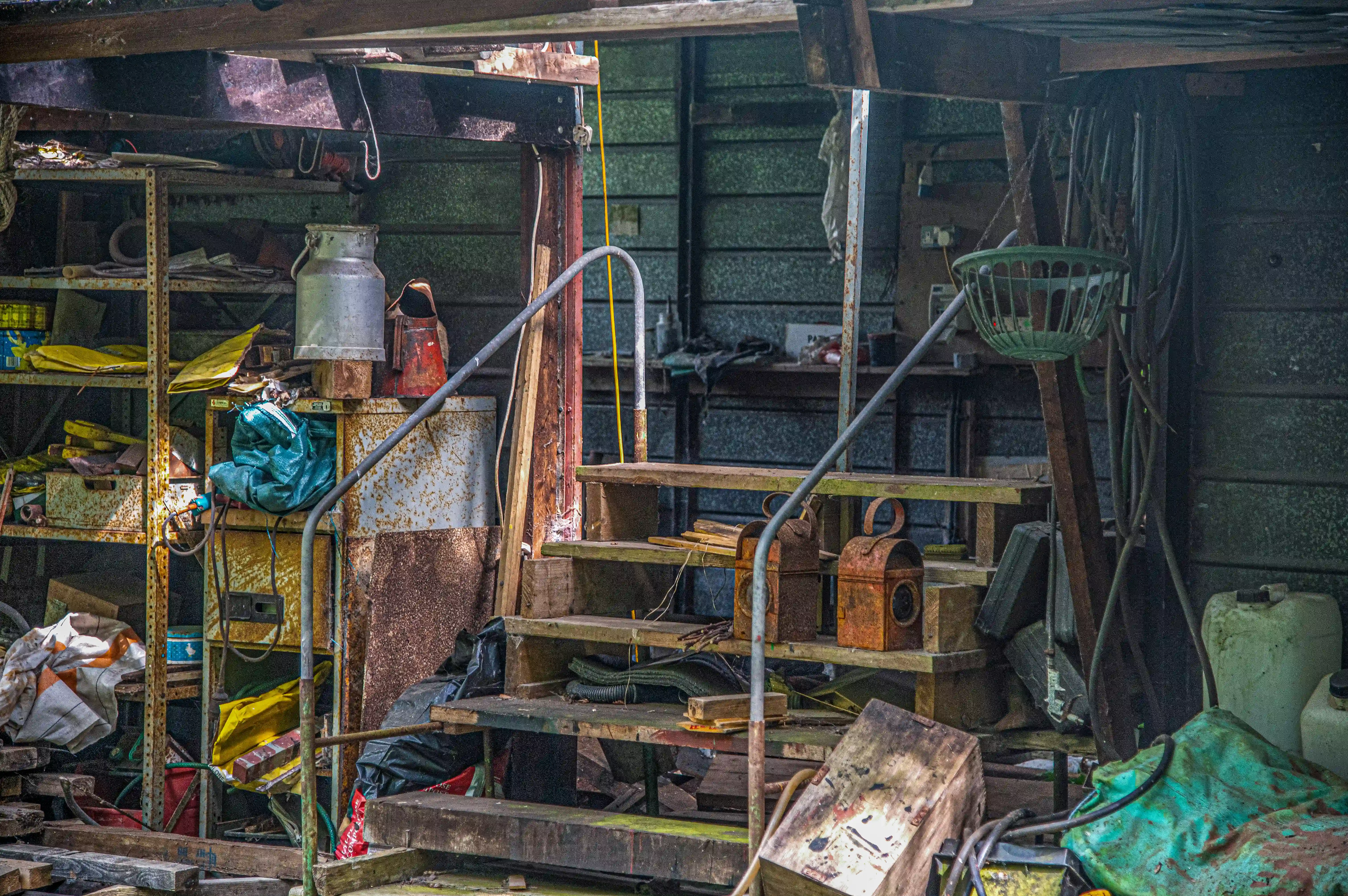 View inside a messy, cluttered shed. Shelves on the left are packed with various containers, tools, and yellow plastic items. Wooden steps lead up into the shed, with two rusty lanterns on the steps. Various tarps, jugs, and hoses are also visible.
