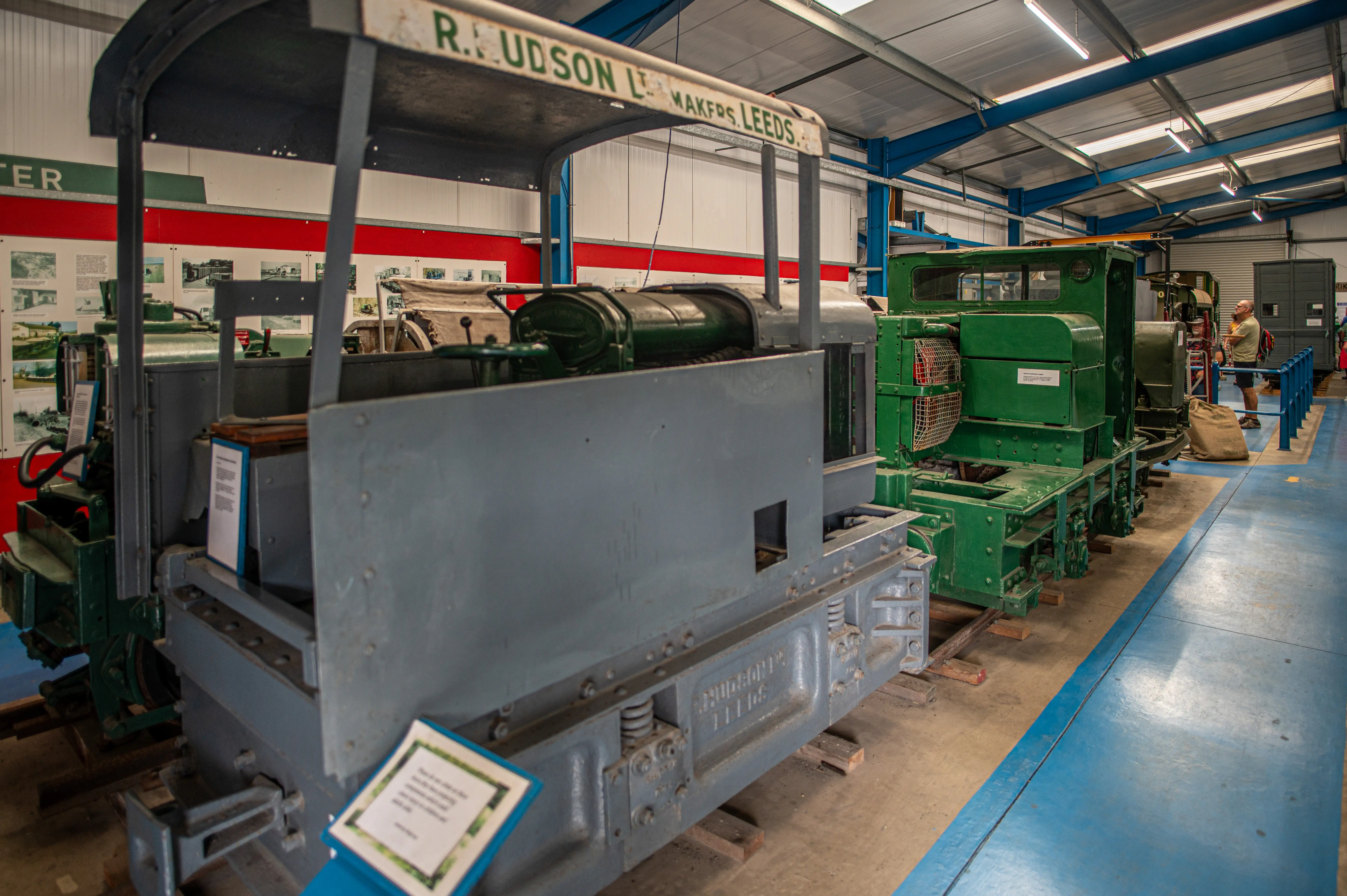 Two narrow-gauge industrial diesel locomotives, a grey one in the foreground and a green one behind it, are parked indoors on a wooden track. The grey locomotive has a maker's plate visible.