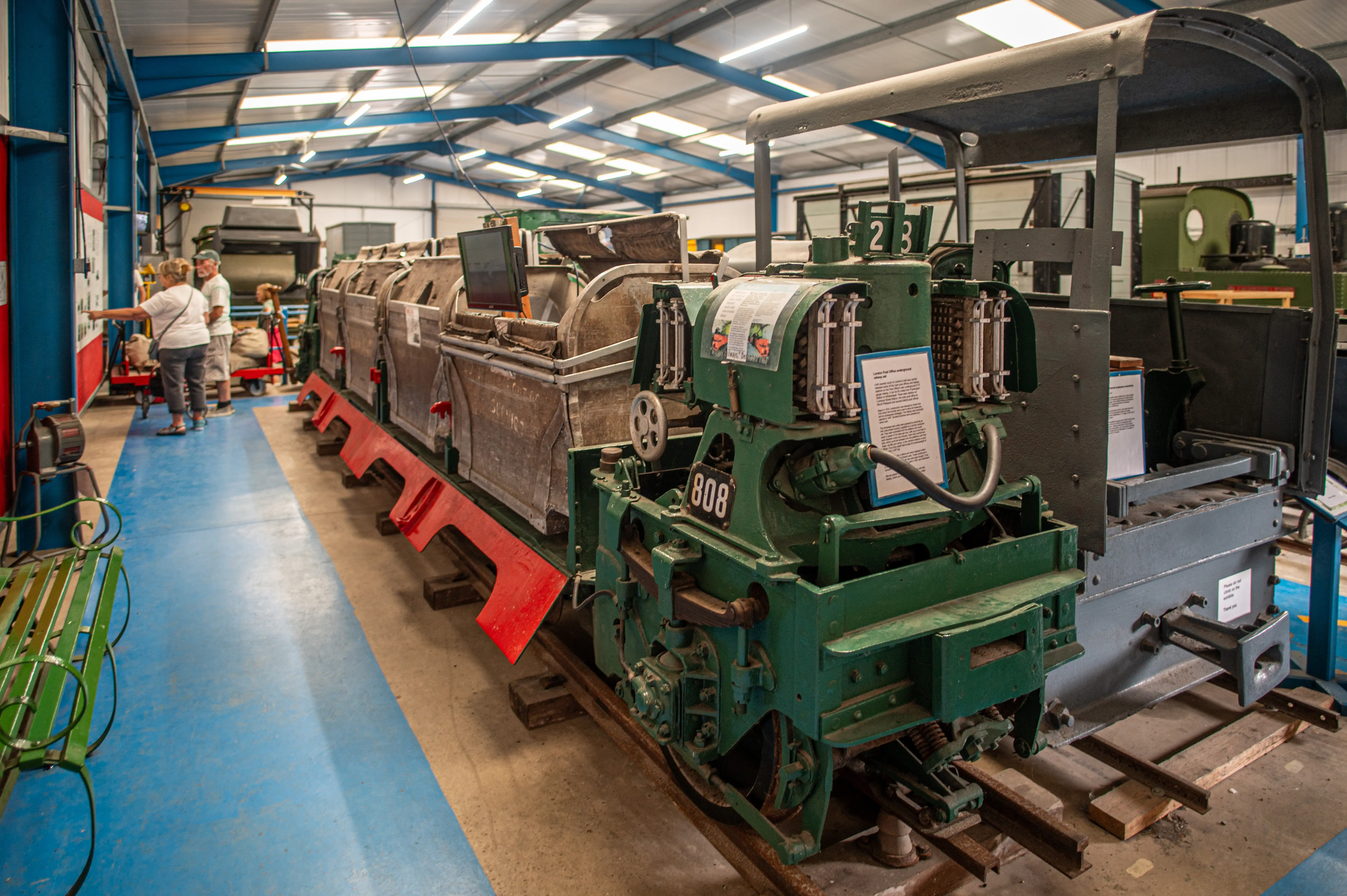 A close-up of a vintage green and grey mail train labelled '808', with two visitors looking at information boards in the background of a museum hall.