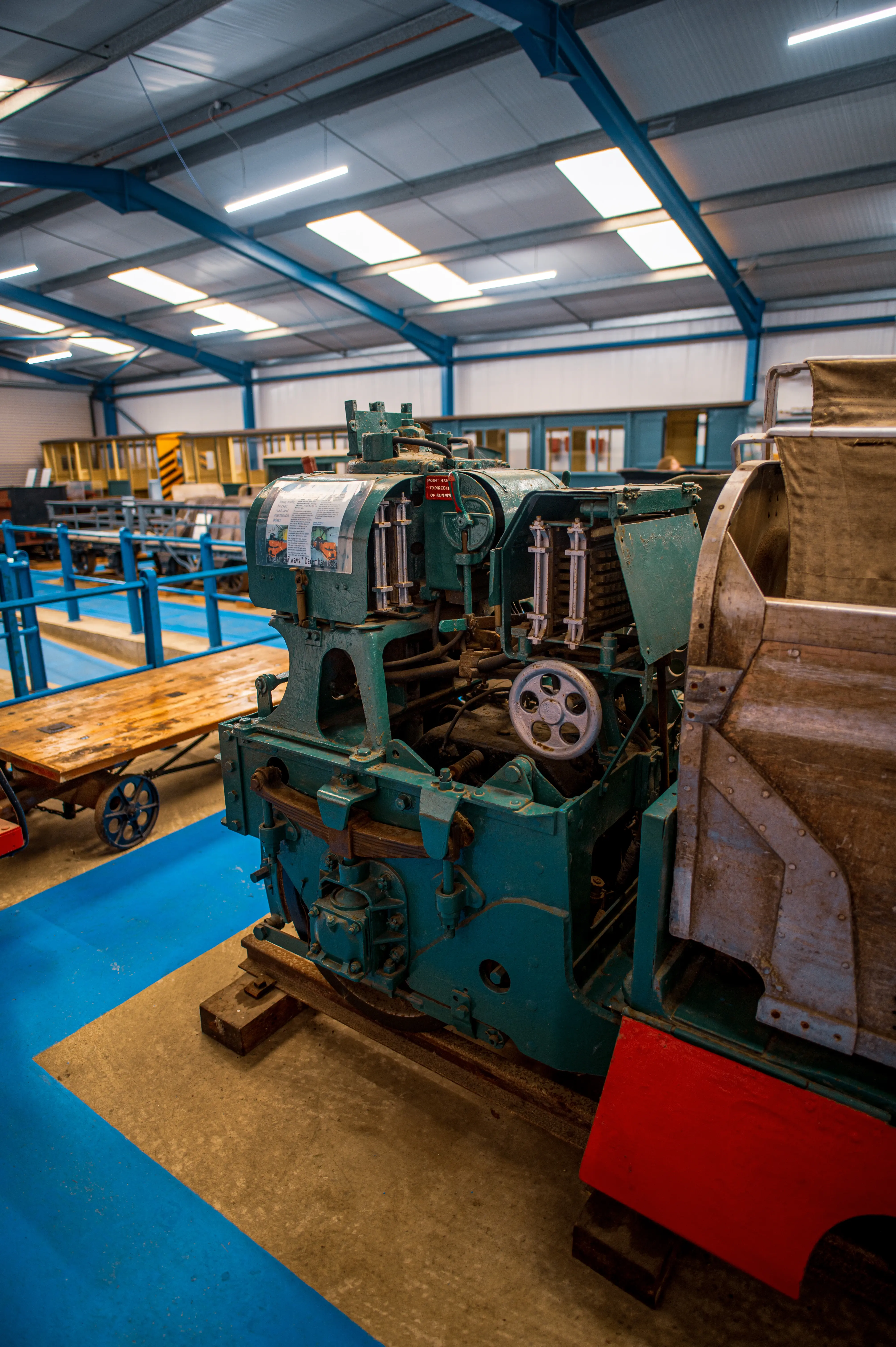 This image captures a detailed section of a steam locomotive's engine, painted in a distinctive teal green, presented in an indoor setting. The machinery exhibits a variety of gears, levers, and structural elements, hinting at the complex operation of historical railway engines. A small informational plaque is affixed to the main unit, suggesting it is part of a museum or heritage railway collection. In the background, other railway carriages and a wooden trolley on wheels can be seen, further contextualising the exhibit within a transportation or industrial history theme. The lighting suggests an exhibition space with overhead fluorescent tubes and skylights. The floor is painted a vibrant blue, with a section of concrete or worn flooring beneath the machinery. The lower front of the exhibit appears to be a red painted section of the locomotive's body.
