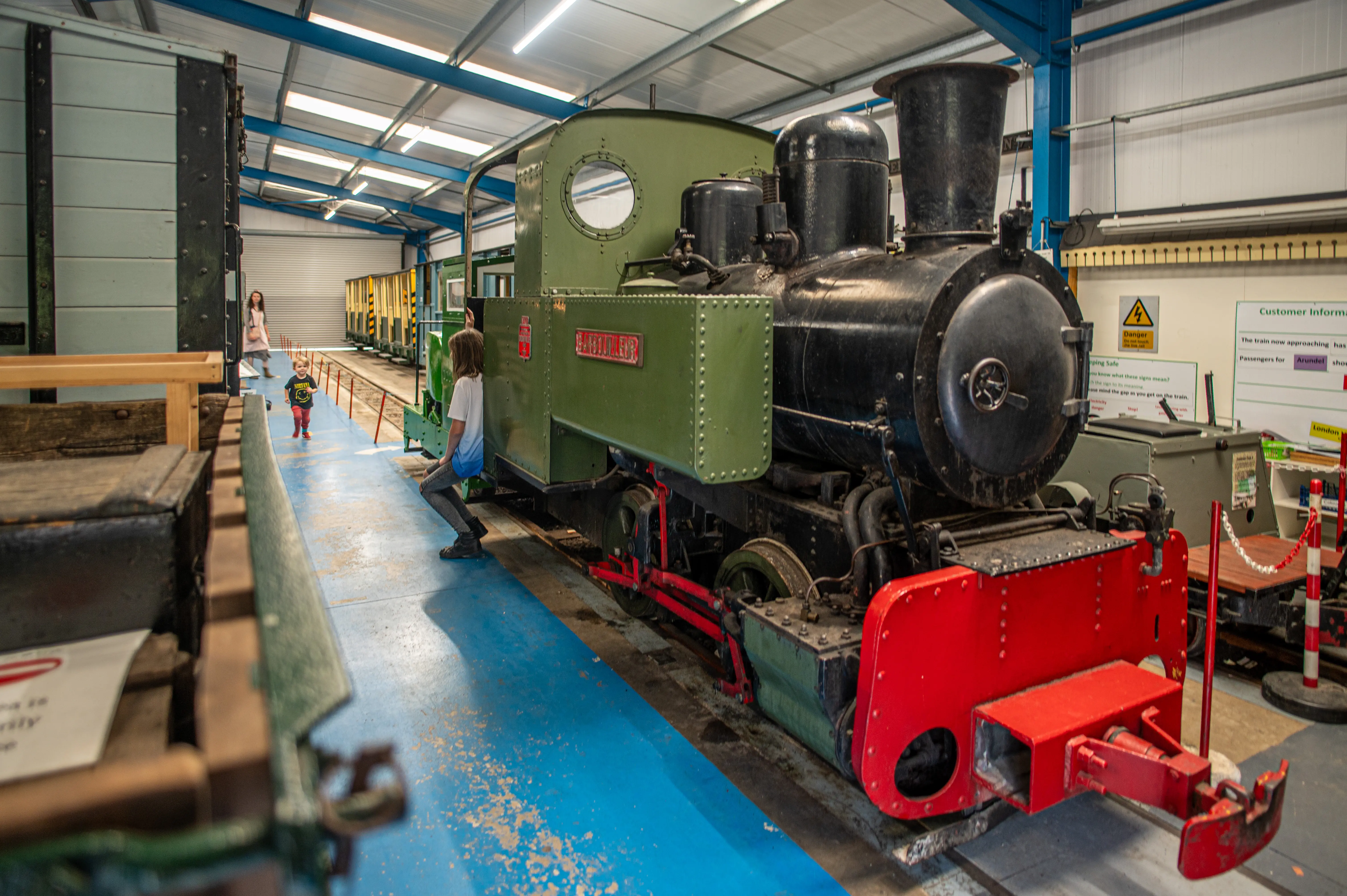 A green and black steam train with a red front is on display in a museum, with two children and an adult nearby on a blue platform.