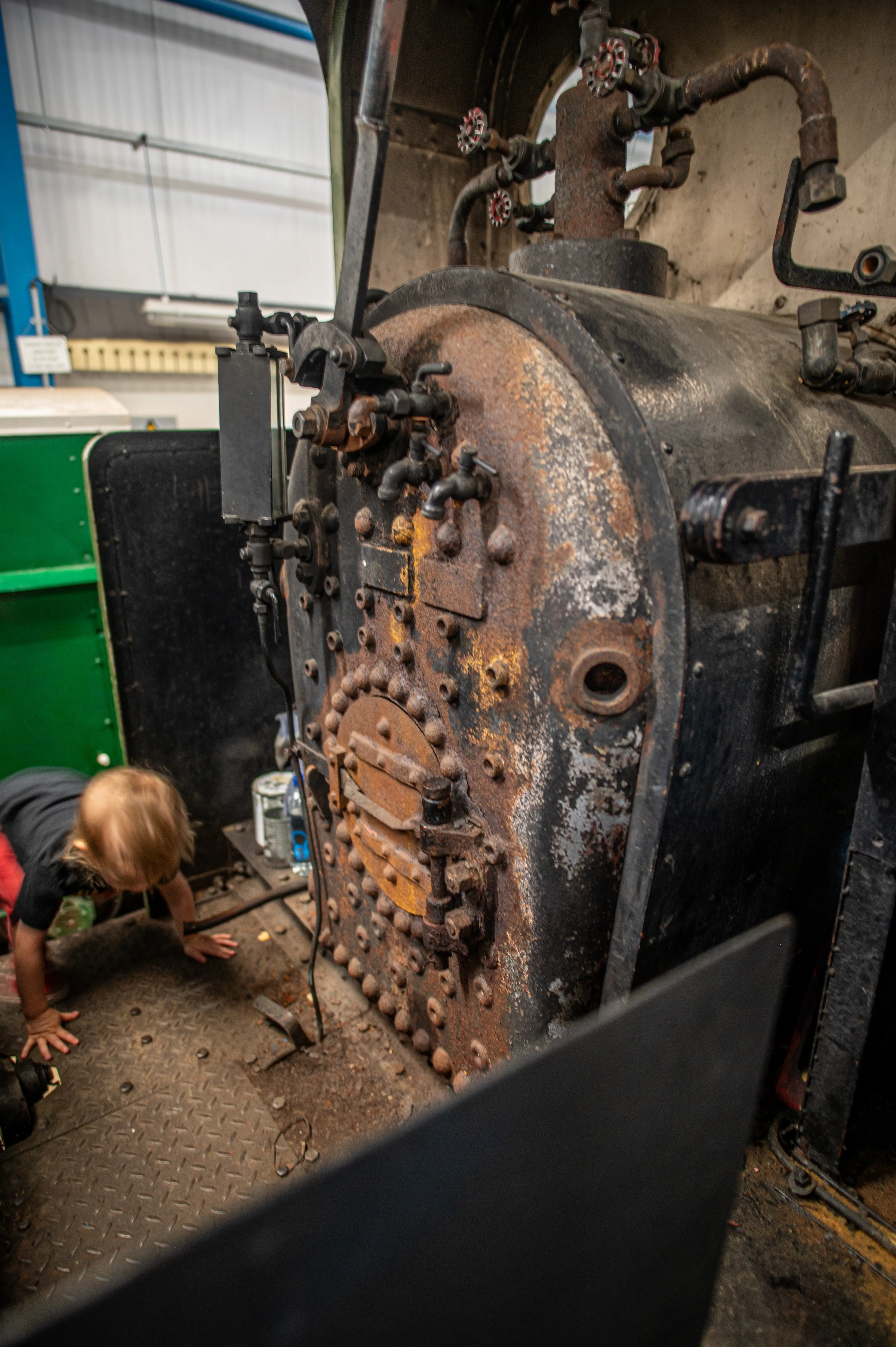 A toddler with blonde hair, wearing a black t-shirt, is on their hands and knees, reaching towards the riveted front of a large, rusty, black steam engine boiler.