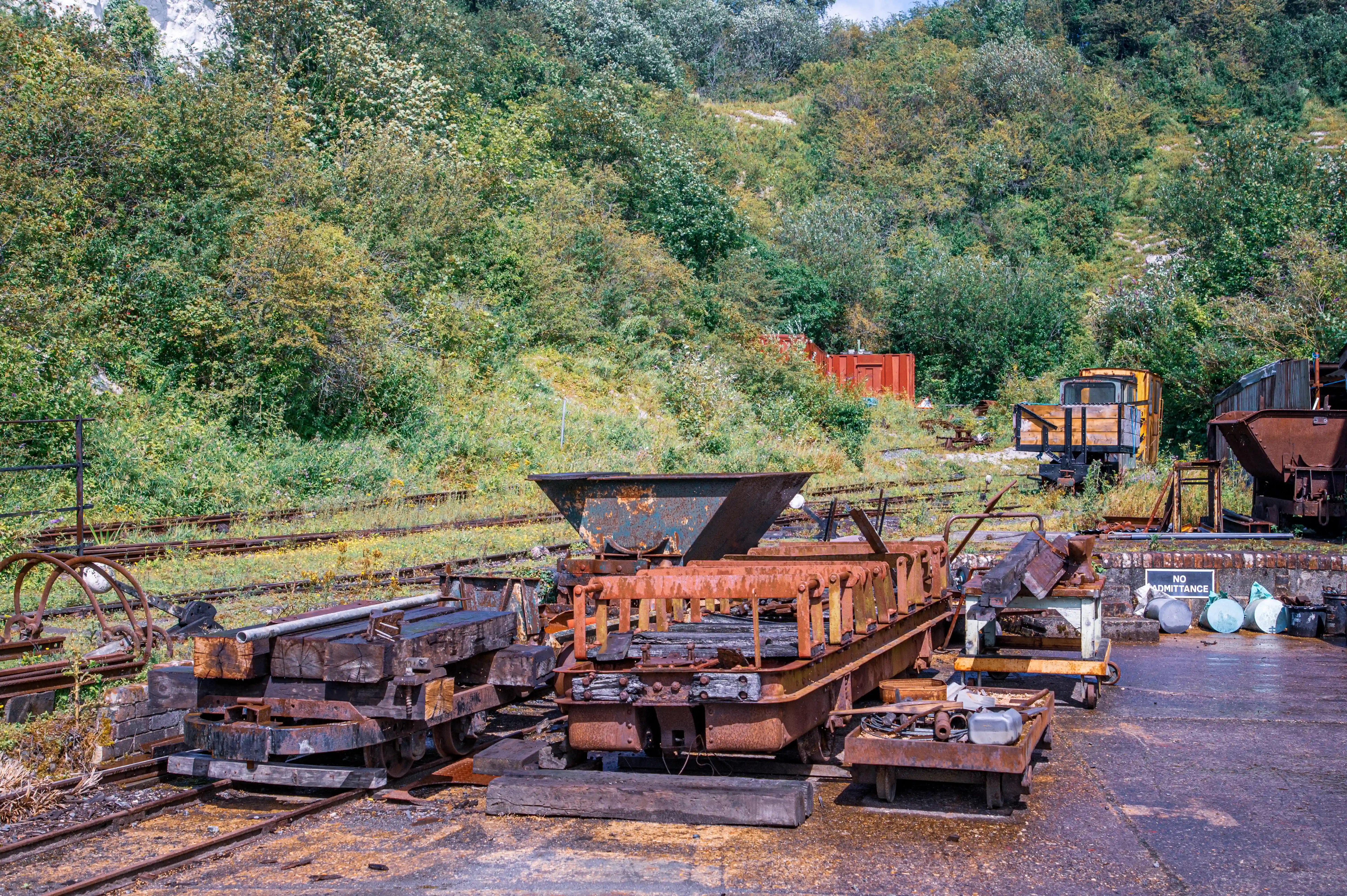Overgrown railway tracks lead past several old, rusty industrial wagons and heavy machinery, with a lush, green hillside rising in the background.