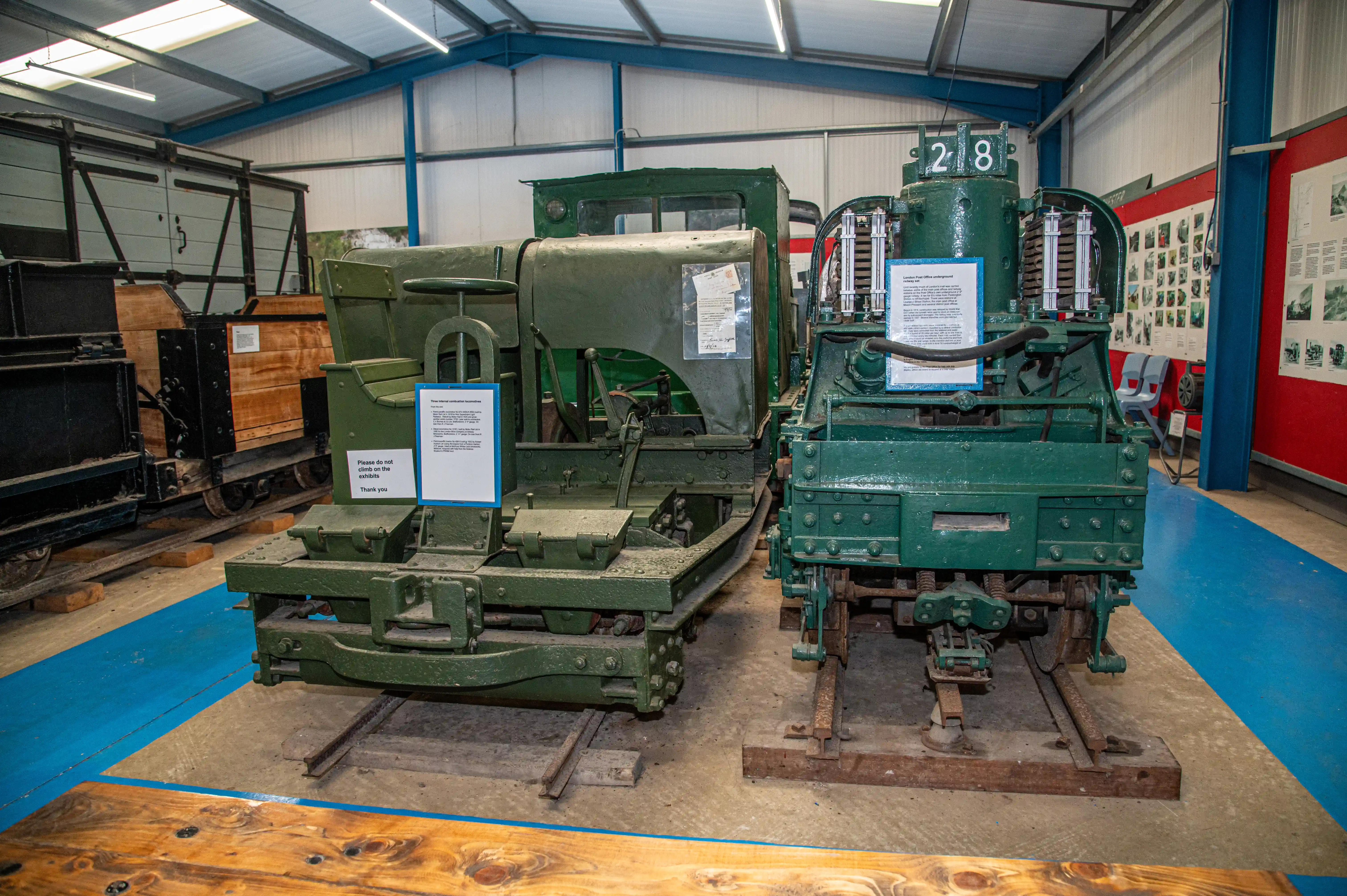 Two green-painted vintage electric shunters are positioned on railway tracks inside a museum. The shunter on the right has the number '28' displayed on its top. Information plaques are visible on both exhibits. Other historical railway items and displays are in the background.