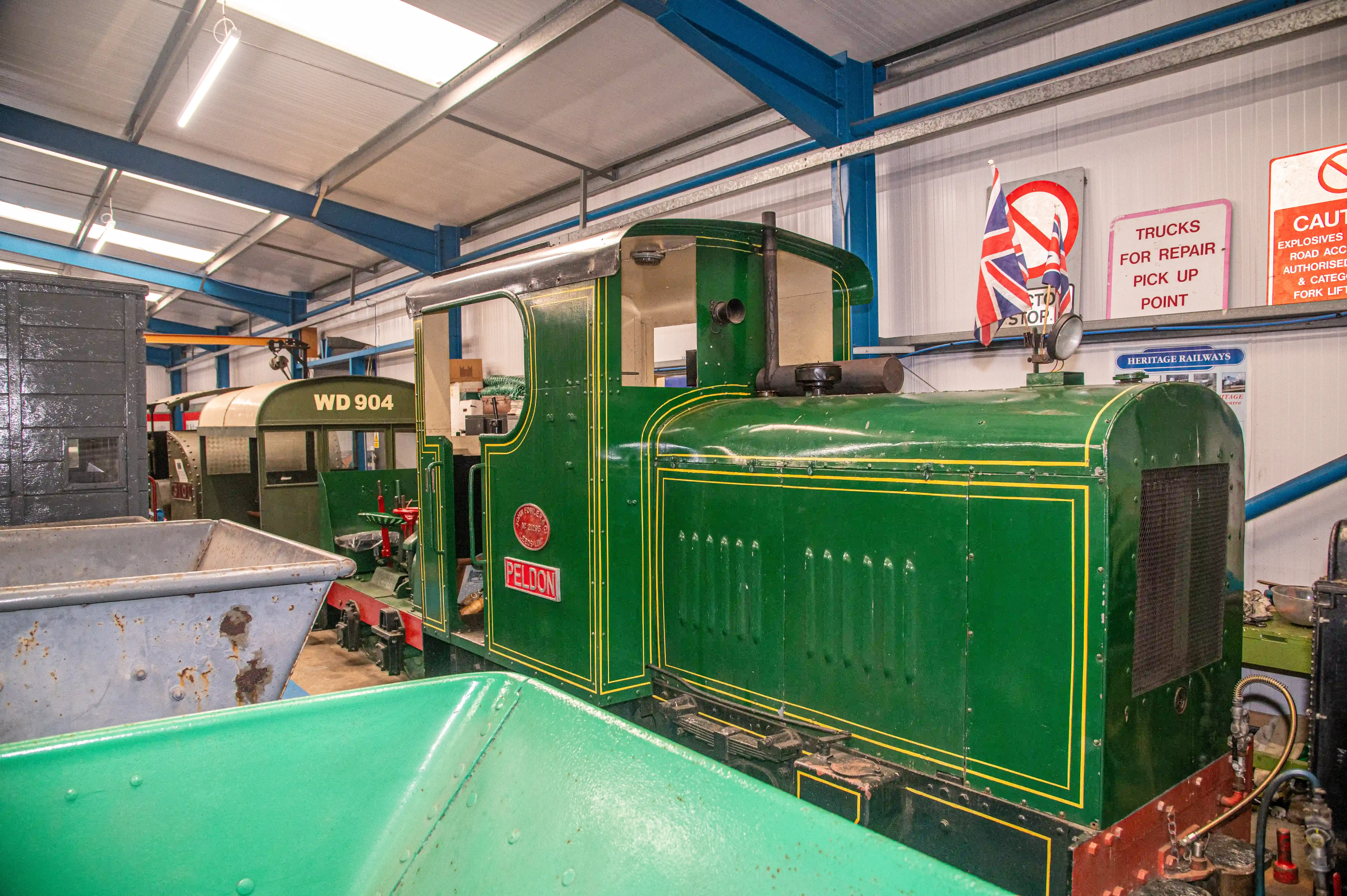 A green heritage steam locomotive named PELDON with yellow trim is parked inside a railway workshop. Another train carriage with the number WD 904 is visible behind it.