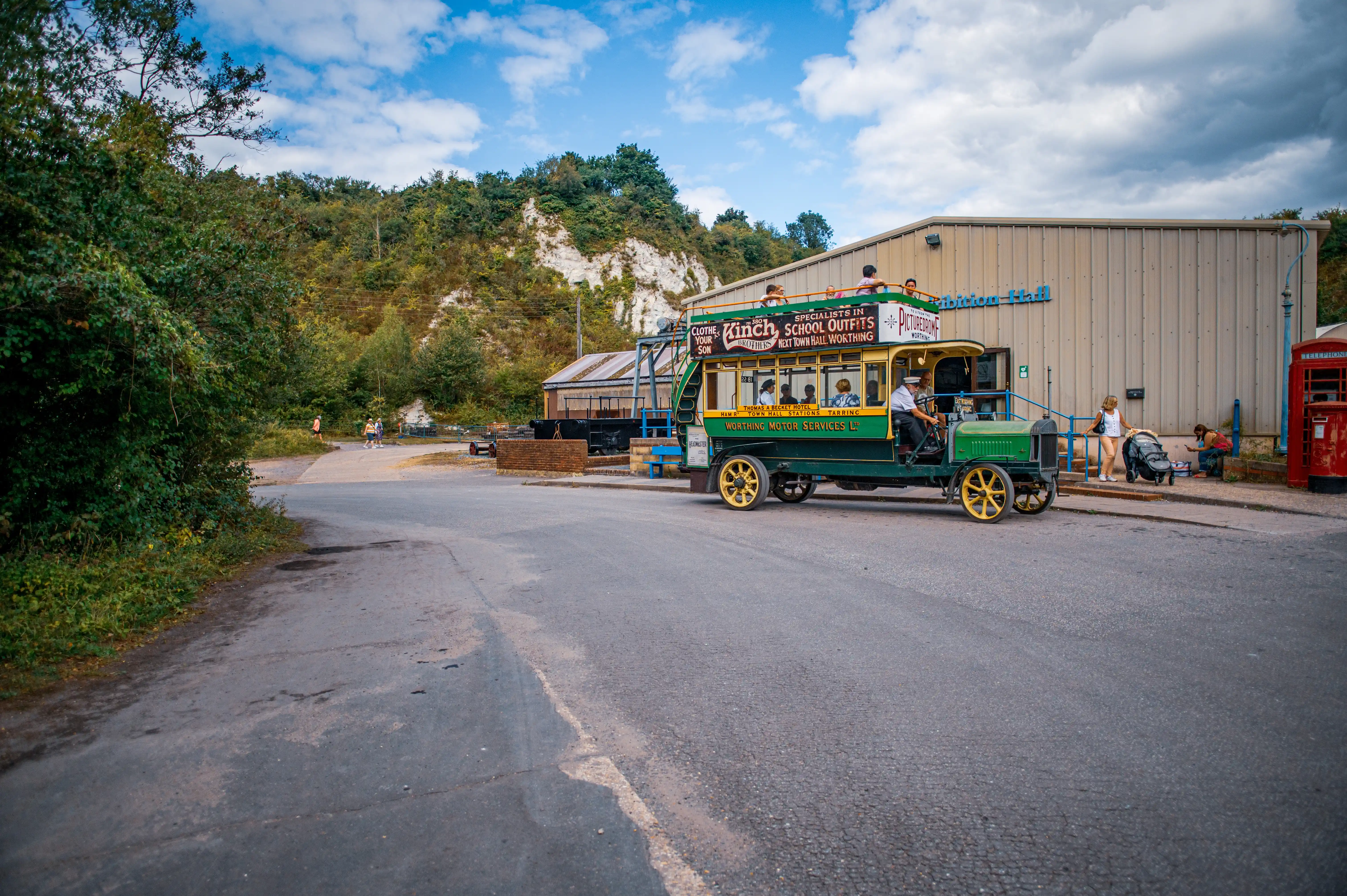 A vintage green and yellow double-decker bus with an open-top upper deck is parked on an asphalt road. The bus has advertisements for 'Kinch School Outfits' and 'Picturedome'. Passengers are visible on the upper deck, and a driver in uniform is at the wheel. Beside the bus is a modern building with ribbed metal siding, labelled 'Exhibition Hall'. A red telephone box stands to the right. In the background, there are trees, a chalky cliff face, and a blue sky with white clouds.