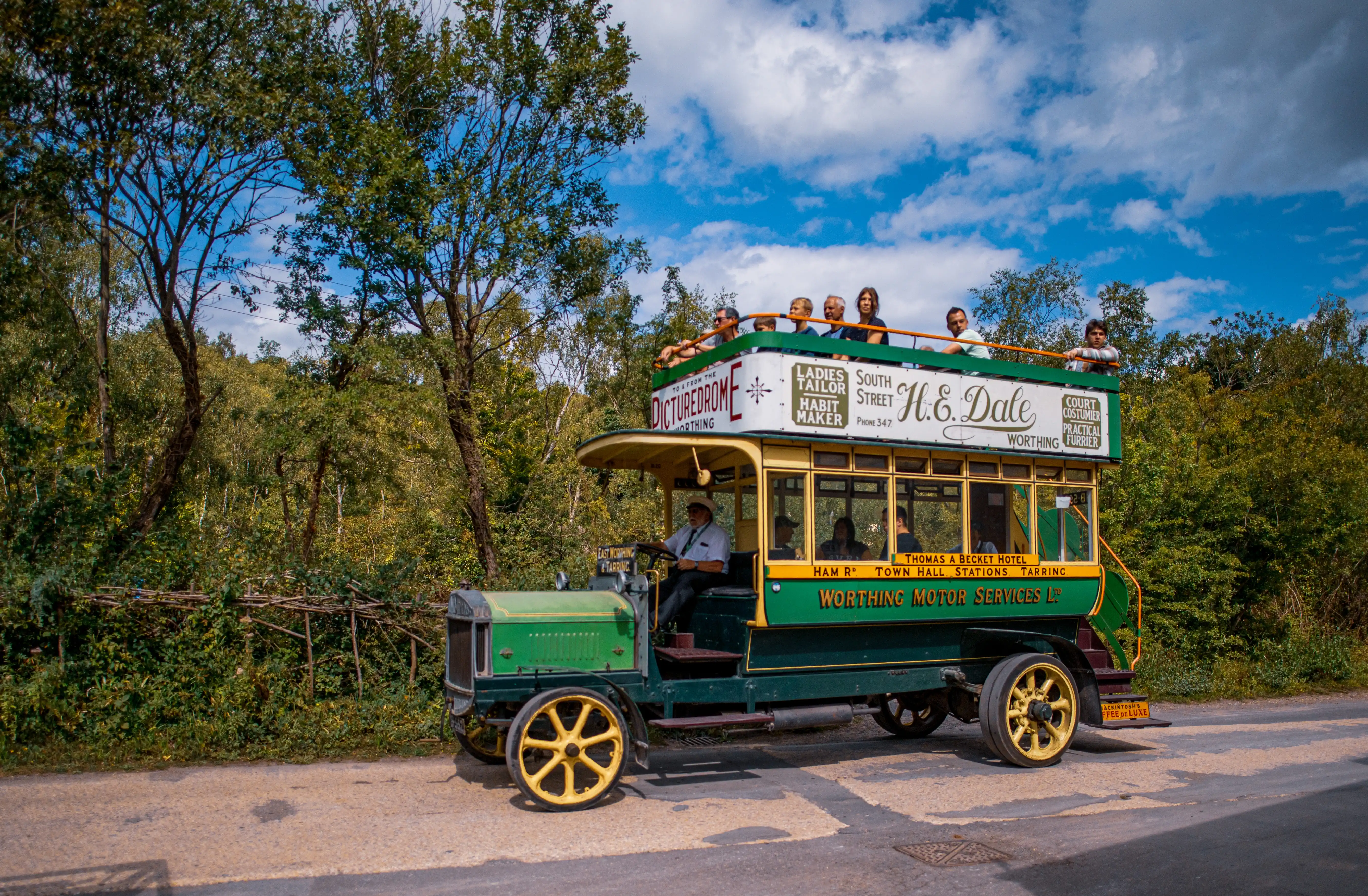 A vintage green and yellow double-decker bus with an open-top upper deck carrying passengers drives along a road bordered by trees under a blue sky.