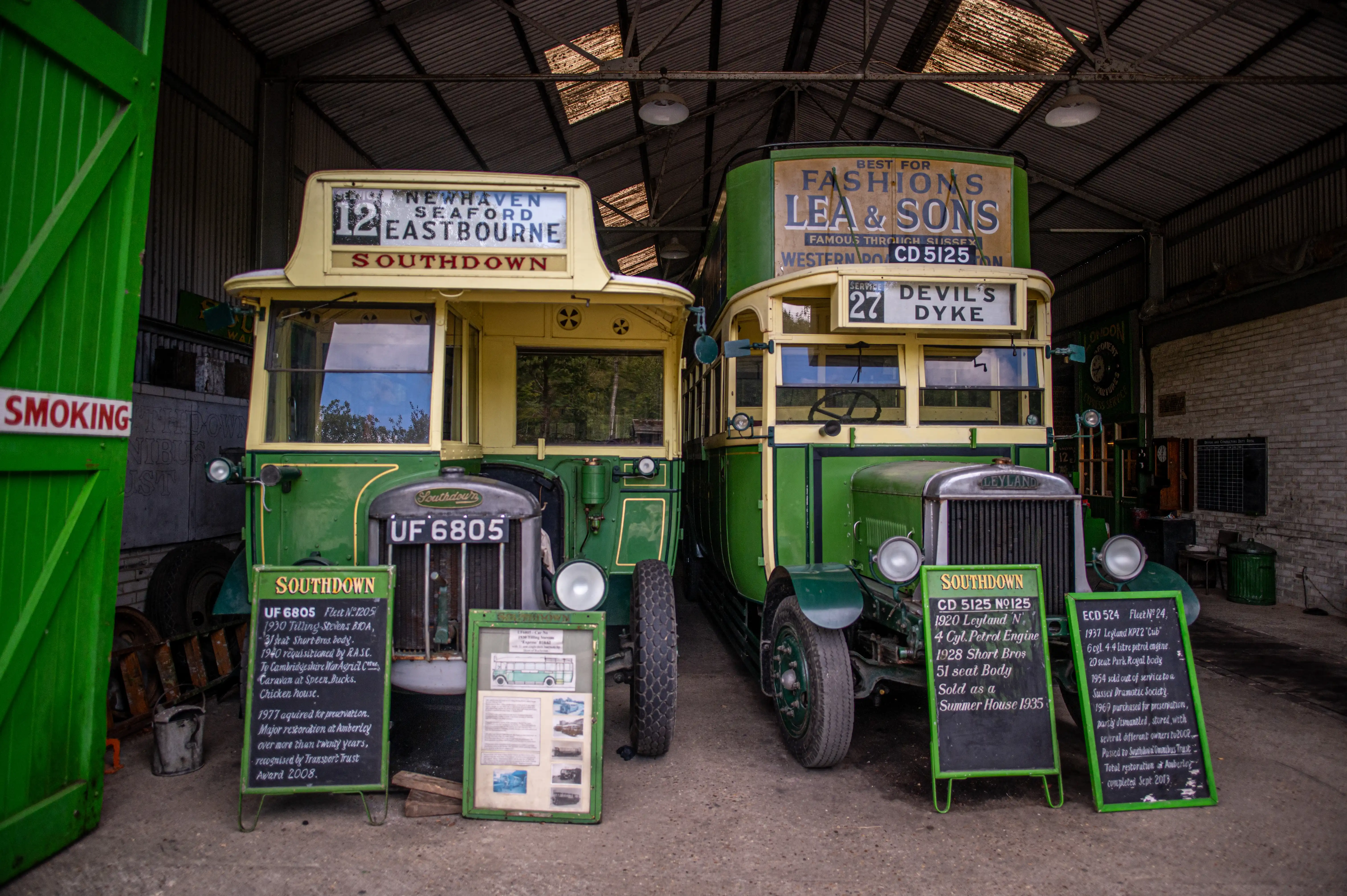 Two vintage green and cream Southdown buses are parked indoors in a Garage. Information boards are in front of each bus, detailing their histories. A 'No Smoking' sign is visible on the left.