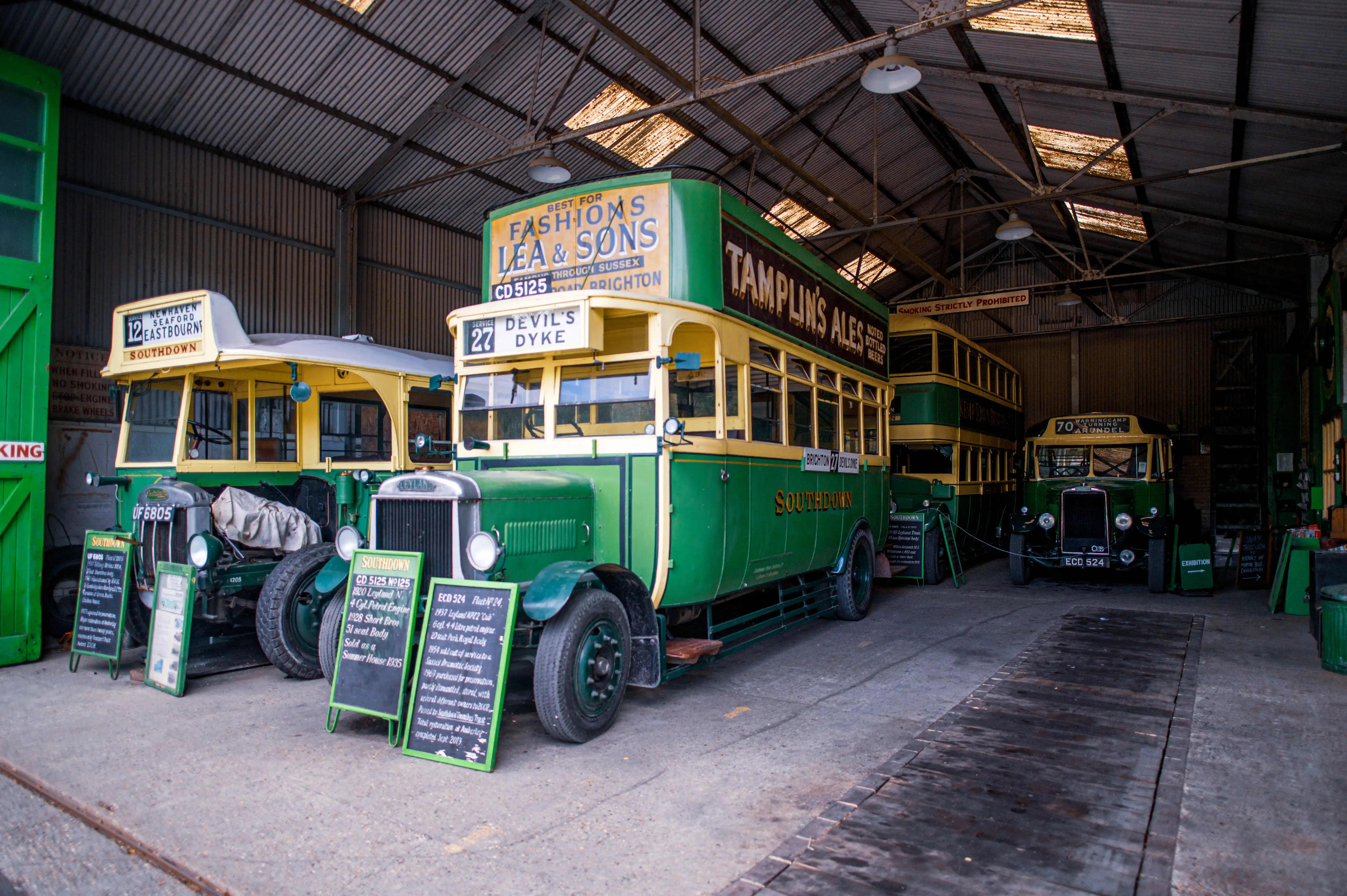Several vintage green and yellow Southdown buses are parked side-by-side in a metal garage. Information boards are placed in front of the buses.