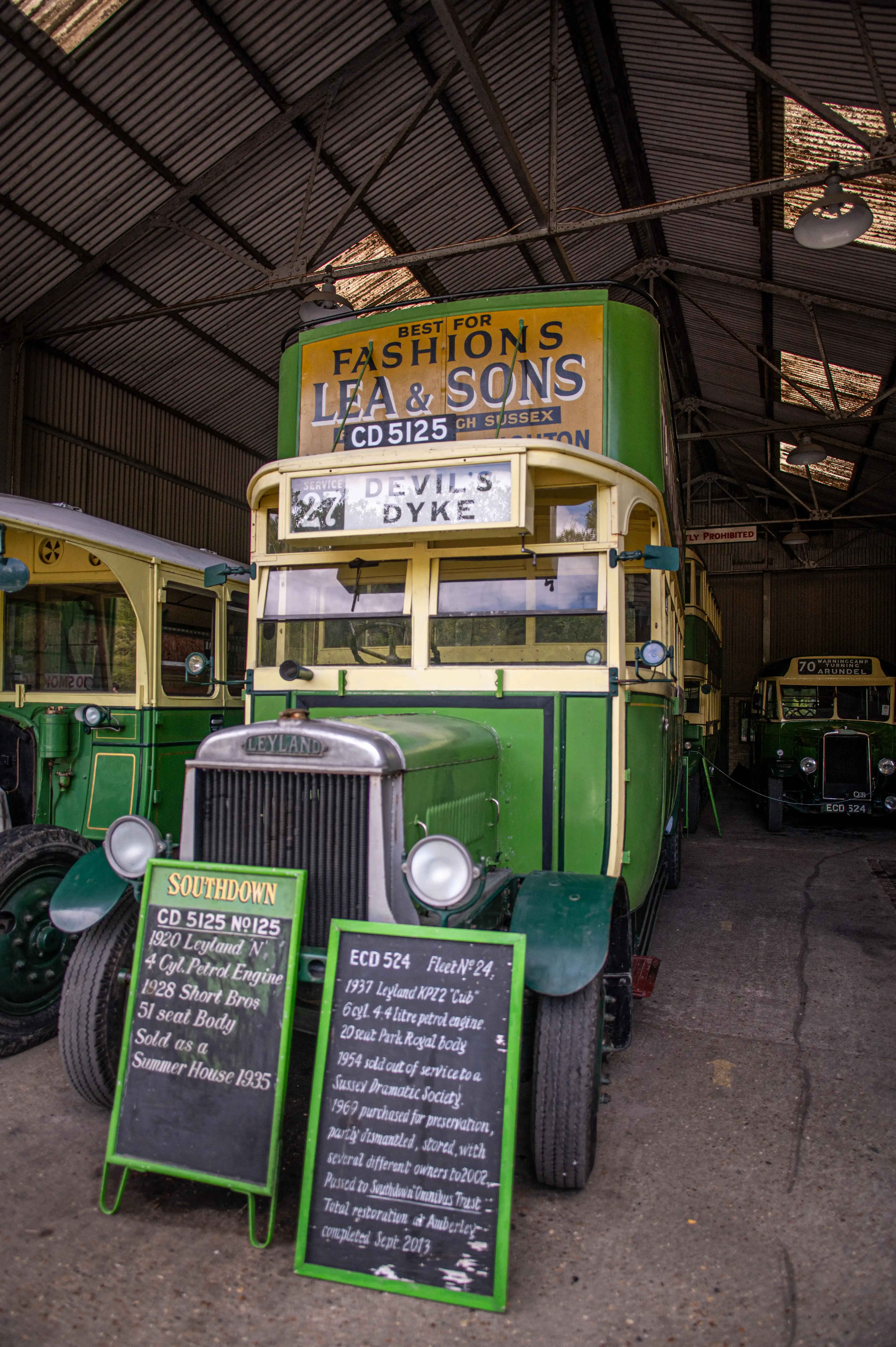 A green and cream double-decker Leyland bus from the Southdown company is parked indoors, with two information boards in front of it detailing its specifications and history. A similar bus is parked behind it, and another one further in the background.